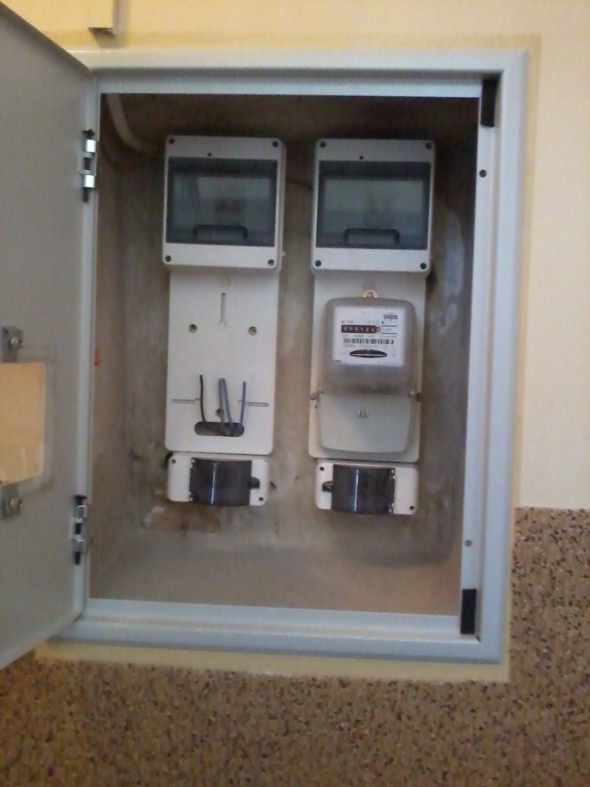 Otwarta skrzynka elektryczna z dwoma licznikami, jeden z wyświetlaczem, drugi przygotowany do montażu, widoczne kable i elementy montażowe.