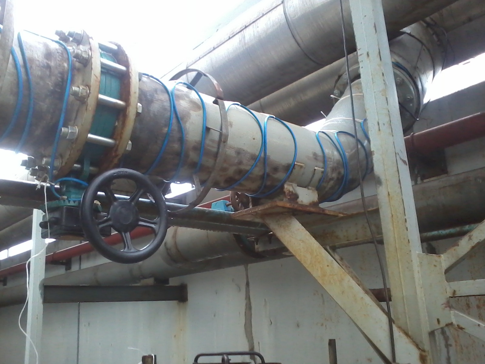 Metalowe rury przemysłowe z zaworem, zabezpieczone niebieską taśmą, zamontowane na stalowej konstrukcji wsporczej.