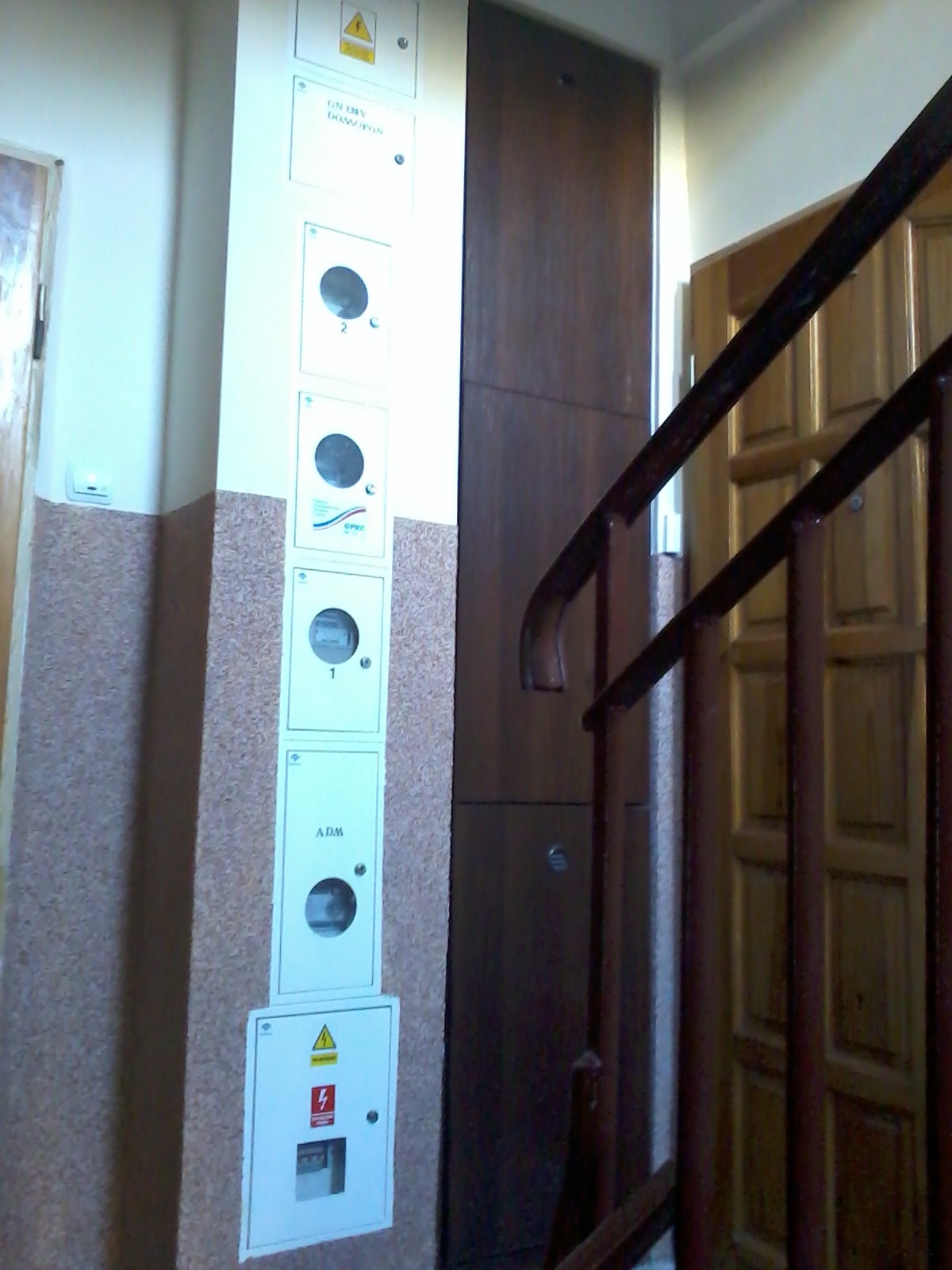 Tablica z licznikami elektrycznymi w klatce schodowej, widoczne skrzynki z oznaczeniami i symbolami ostrzegawczymi, ujęcie z perspektywy schodów.