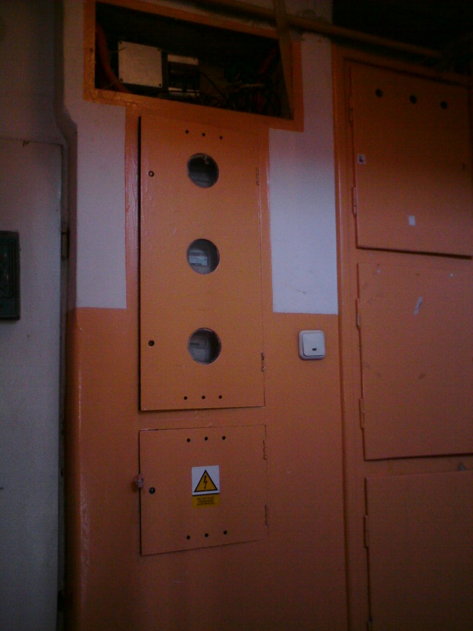 Pomarańczowa skrzynka rozdzielcza z trzema okrągłymi otworami i znakiem ostrzegawczym o napięciu, widoczne przewody w górnej części, słabe oświetlenie