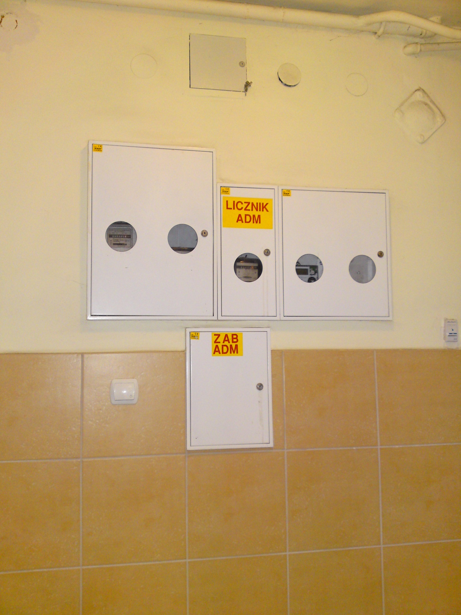 Zestaw białych skrzynek licznikowych z żółtymi etykietami 'Licznik ADM' i 'ZAB ADM' zamontowanych na ścianie nad brązowymi płytkami, widoczne liczniki przez okrągłe otwory.