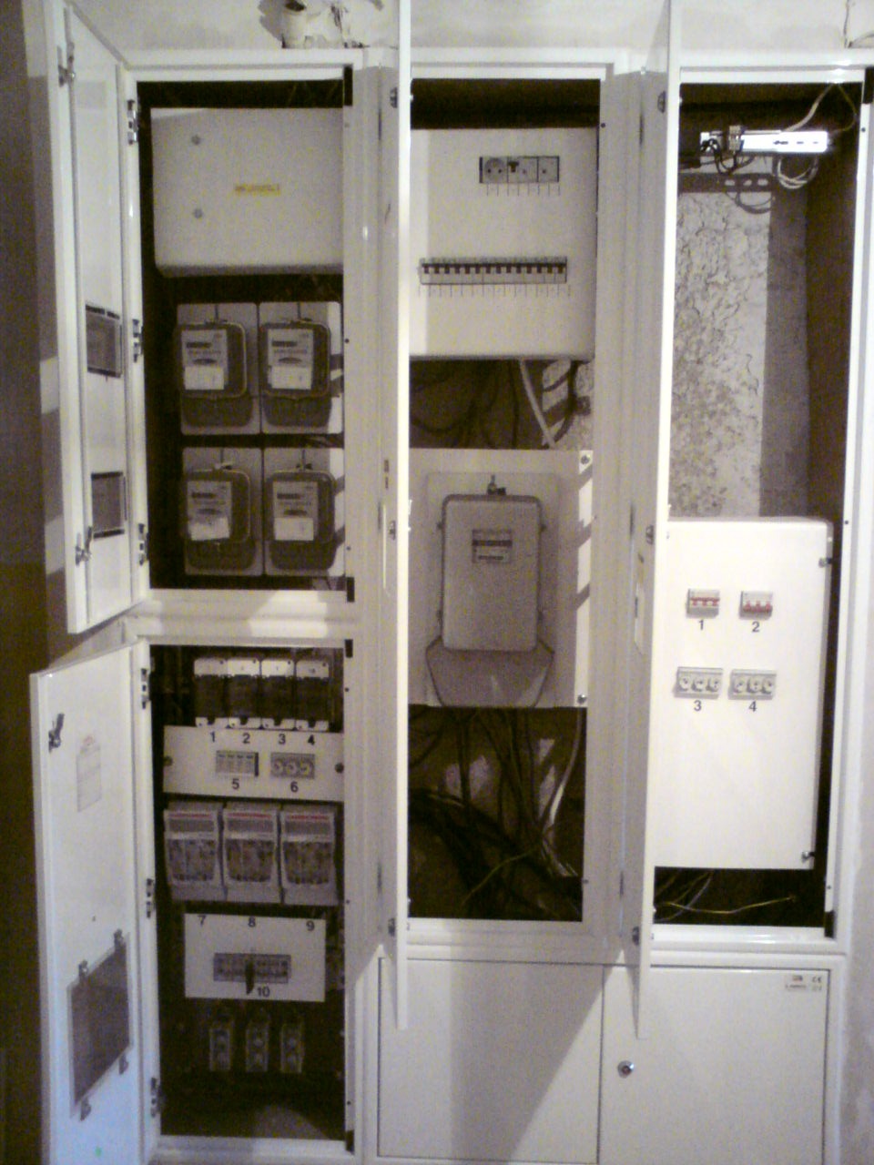 Otwarta szafa rozdzielcza z licznikami elektrycznymi, bezpiecznikami i okablowaniem wewnątrz, widoczne oznaczenia numeracyjne na panelach.