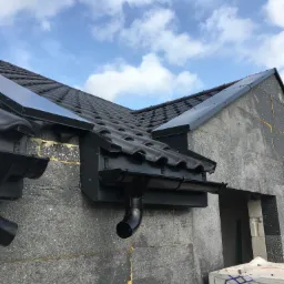 Fragment domu w trakcie budowy z widocznym dachem z czarnej dachówki, orynnowaniem i szarą ścianą z bloczków betonowych oraz częściowo widocznym niebem.