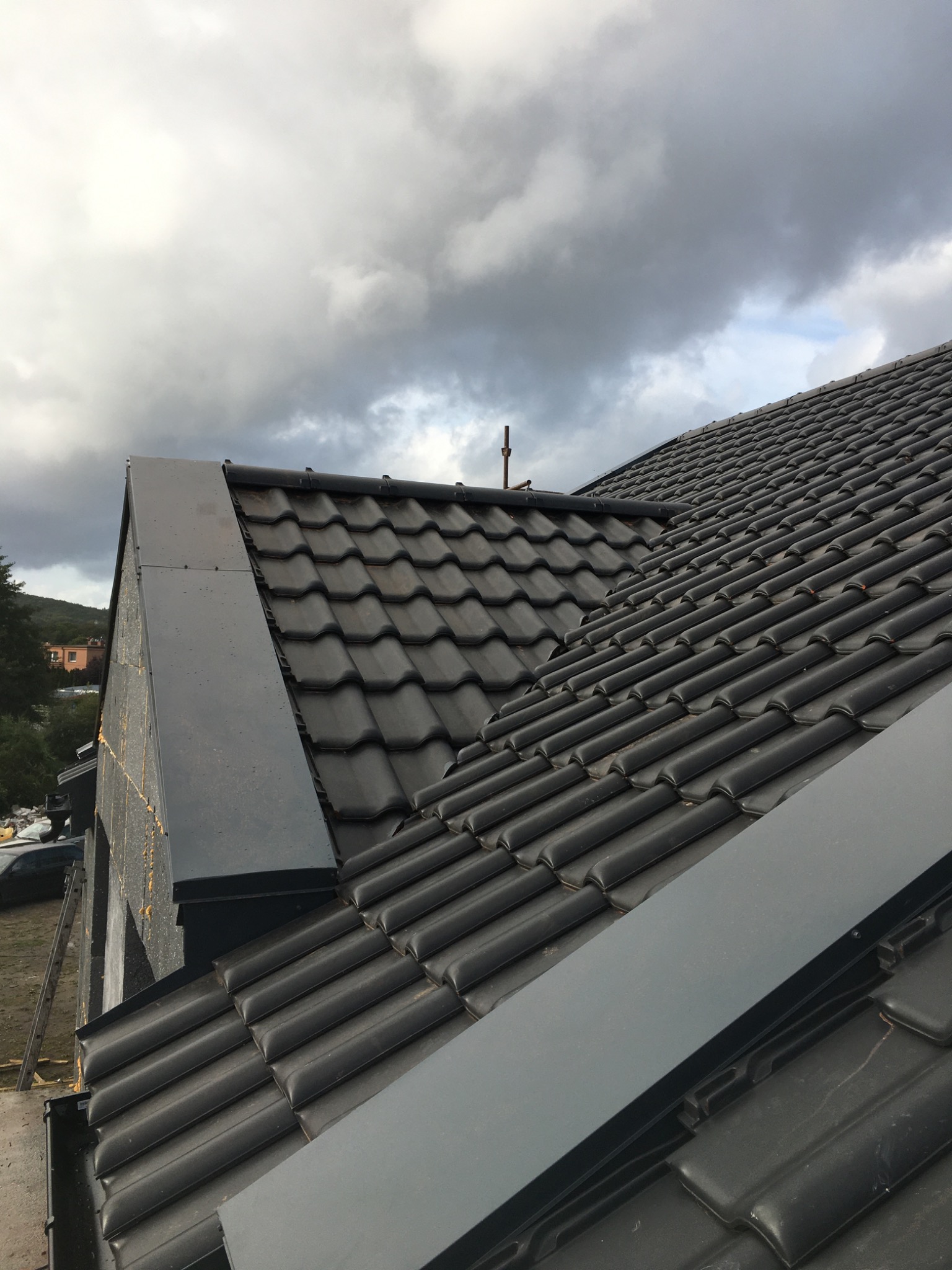 Czarny dach z dachówki ceramicznej, z widocznymi obróbkami blacharskimi, wykonany w trakcie budowy domu, pochmurne niebo w tle.