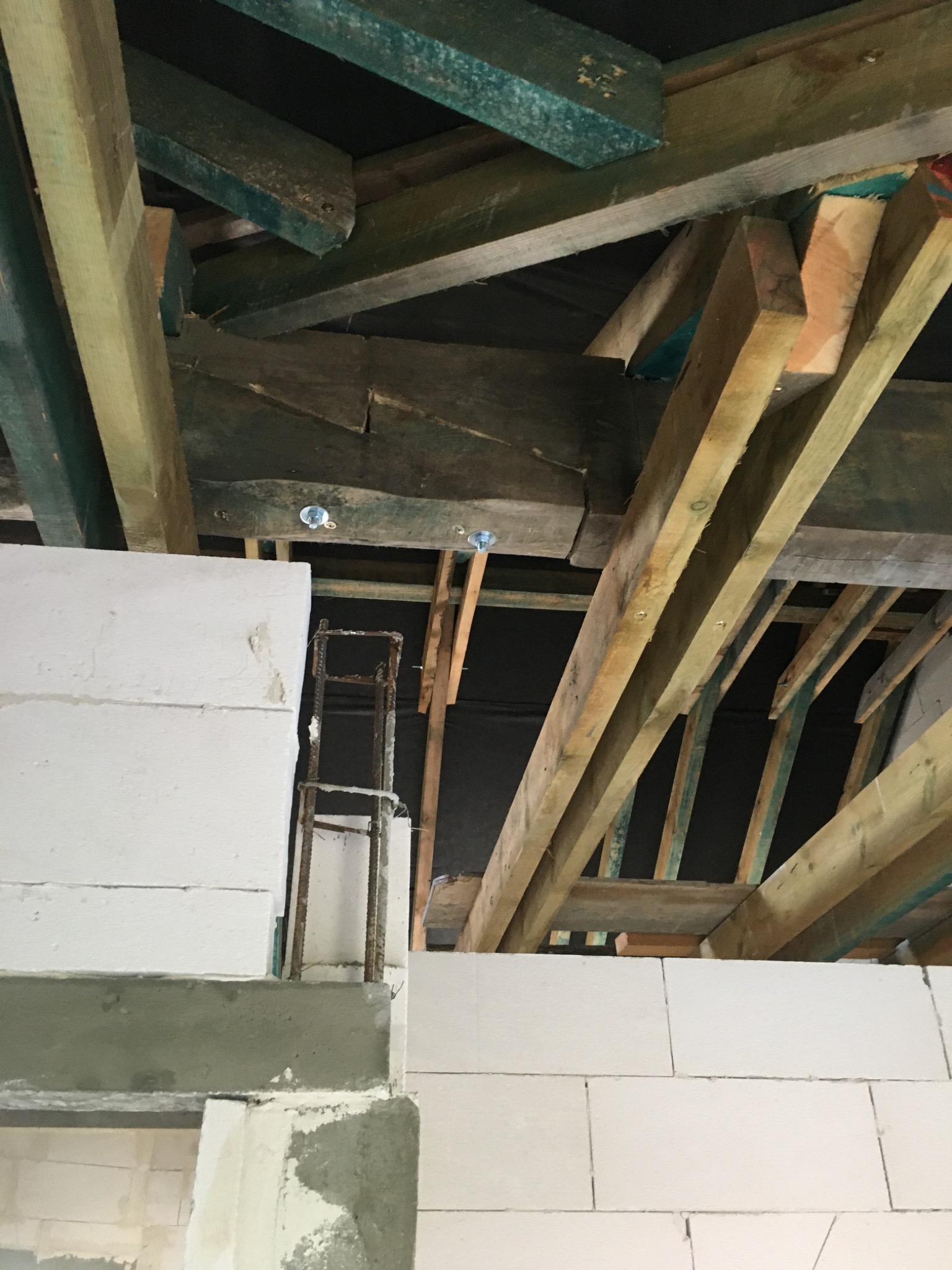 Wnętrze budowy w Gliwicach: Ściana z białych bloczków, zbrojenie i drewniana konstrukcja dachu w trakcie montażu, widoczne elementy mocujące.