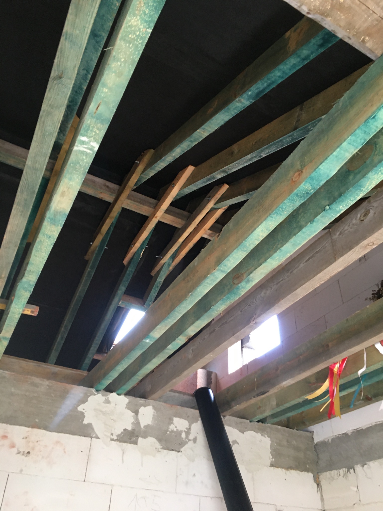 Wnętrze konstrukcji dachu z drewnianymi belkami pomalowanymi na zielono-niebieski kolor, widoczne fragmenty ścian z białego betonu komórkowego i czarna rura wentylacyjna.