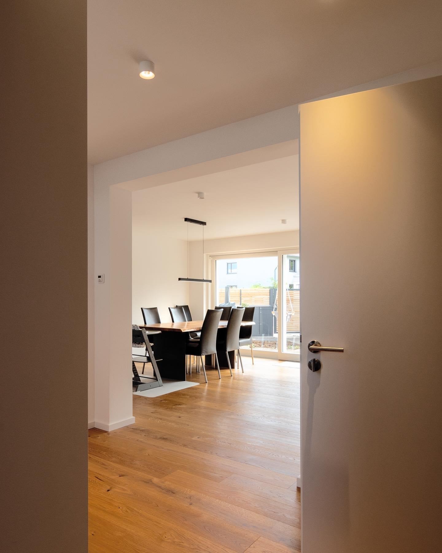 Widok na jasną jadalnię z drewnianą podłogą i stołem z czarnymi krzesłami przez częściowo otwarte, białe drzwi z metalową klamką.