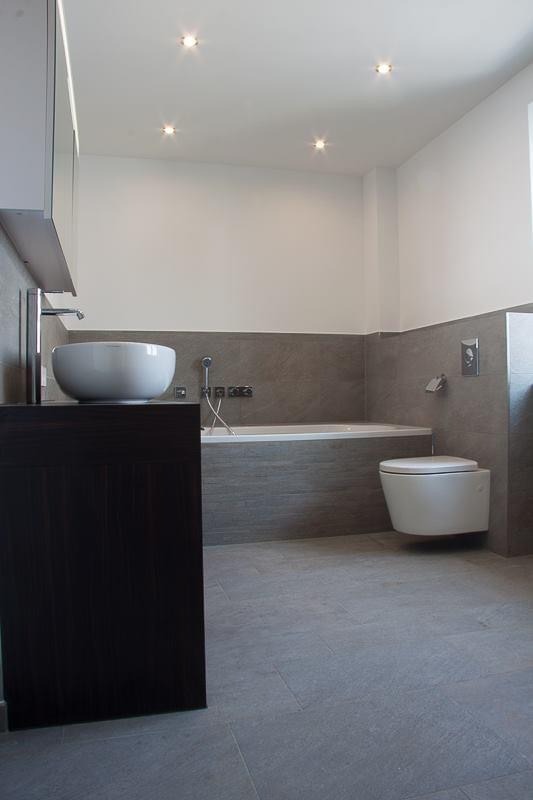 Nowoczesna łazienka z szarą płytką, wiszącą toaletą i okrągłą umywalką na ciemnym blacie.