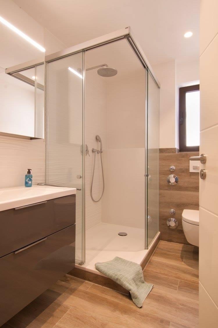 Nowoczesna łazienka z kabiną prysznicową typu walk-in, umywalką z szafką w kolorze taupe, drewnopodobną podłogą i akcentami w kolorze drewna na ścianie oraz zielonym ręcznikiem na podłodze.