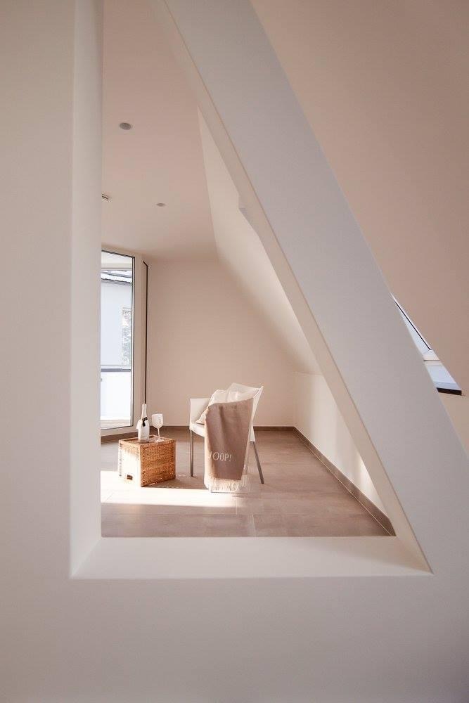 Jasne wnętrze poddasza z oknem, skośnym sufitem i beżowymi płytkami podłogowymi. Na krześle leży beżowy pled z napisem Joop!, a obok stoi wiklinowy stolik z butelką i kieliszkami.