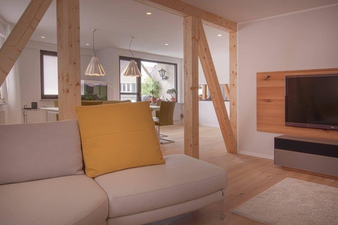 Jasne wnętrze salonu z drewnianymi belkami konstrukcyjnymi, widok z kanapy na jadalnię i okno z roślinami, telewizor na drewnianej ścianie.