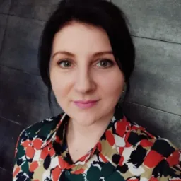 Portret kobiety z ciemnymi włosami, zielonymi oczami i różowymi ustami, ubrana w kolorową koszulę w kwiaty na tle szarej ściany z płytek.