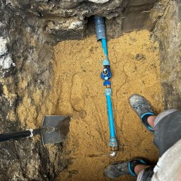 PLOOM USŁUGI HYDRAULICZNE PRZEMYSŁAW KUBIAK - Wykop z piaskiem, w którym widoczna jest pionowa, niebieska rura wodna z zaworem, częściowo zamontowana w ścianie wykopu, obok łopata i noga w roboczym obuwiu.