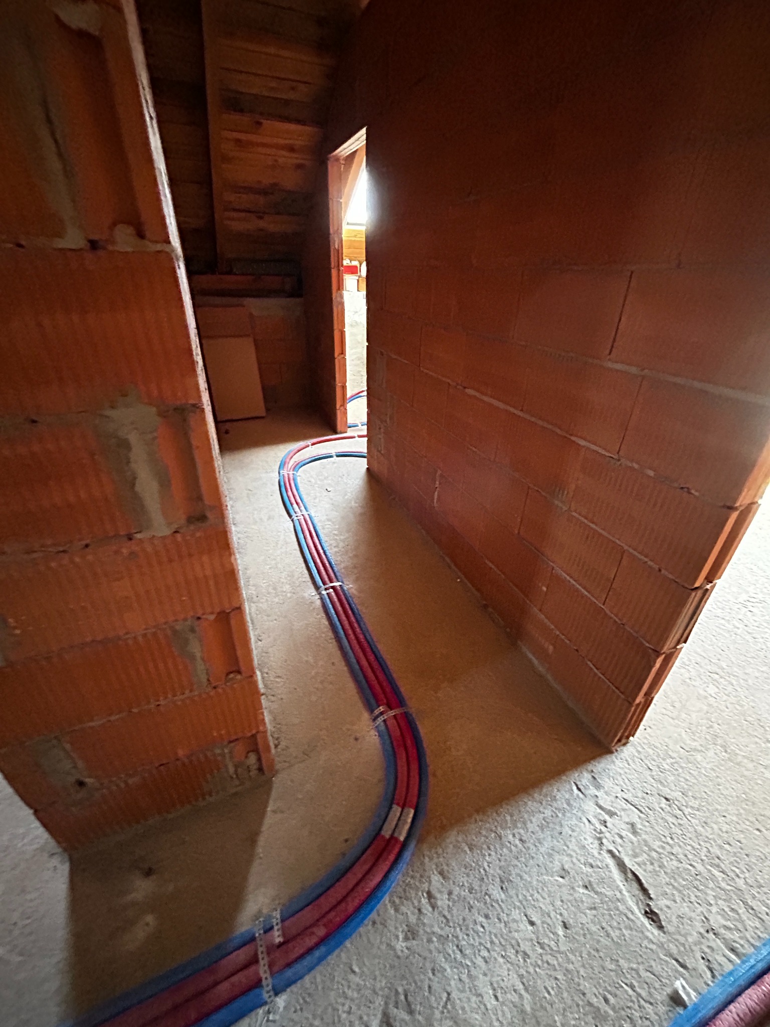 Wnętrze w trakcie budowy: czerwono-niebieskie rury instalacyjne prowadzone wzdłuż surowych ścian z cegły ceramicznej, widoczne mocowania do podłoża.