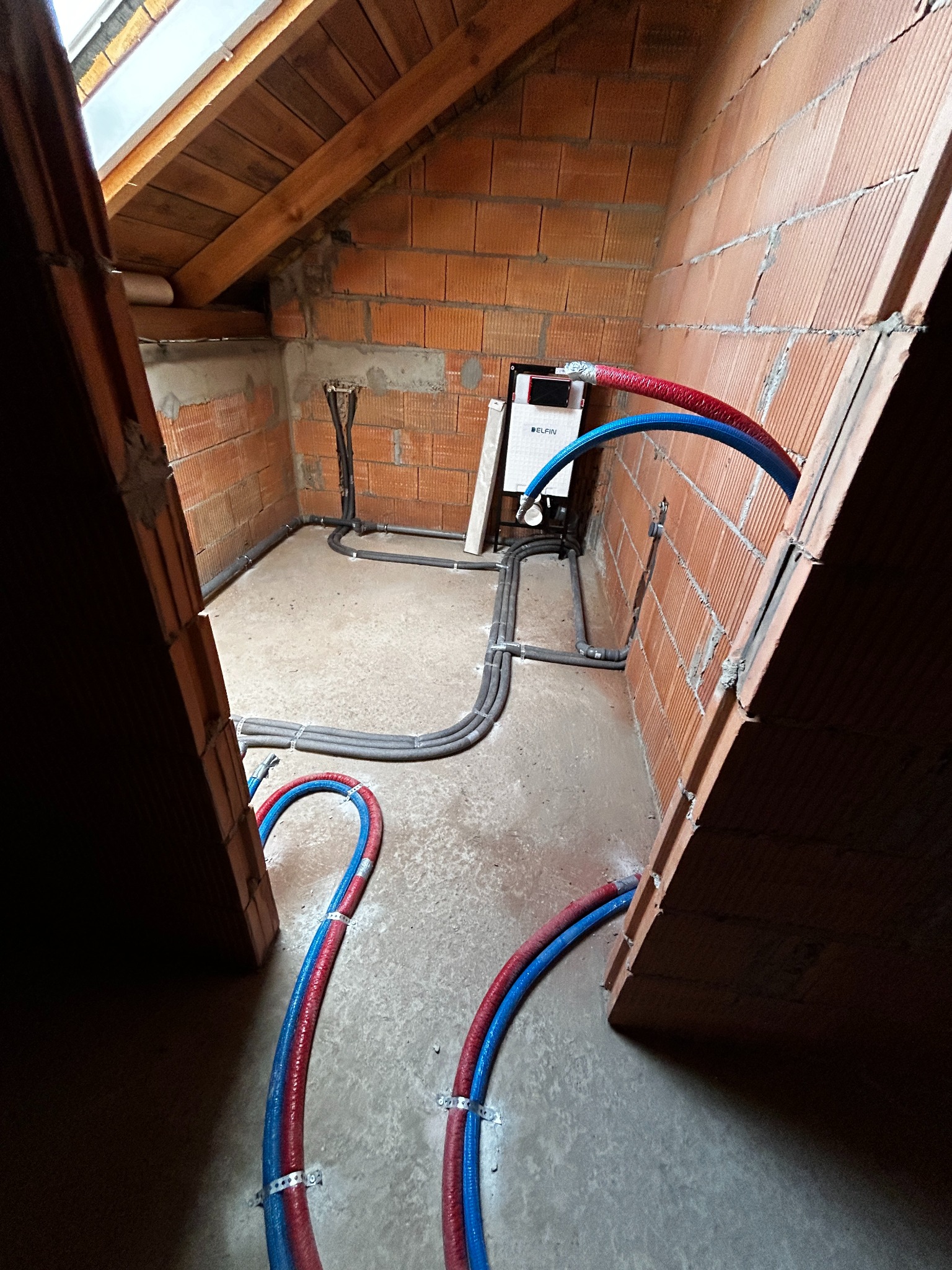 Surowy stan poddasza z widoczną instalacją grzewczą: rury w izolacji czerwono-niebieskiej przymocowane do ściany z pustaków, szare rury instalacji rozprowadzone po podłodze, biały piec na ścianie...