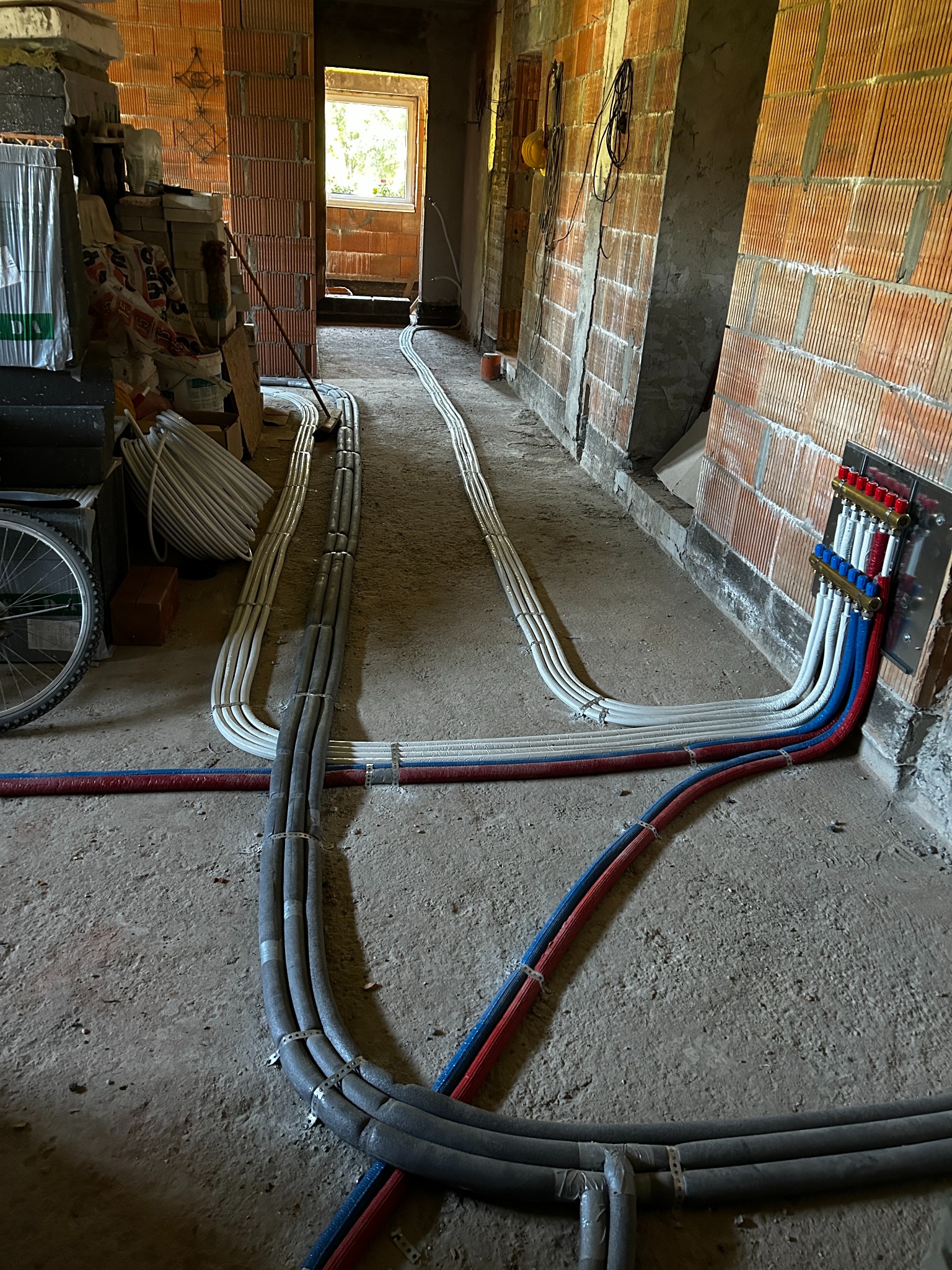 Rozprowadzenie rur instalacji grzewczej i wodnej w surowym stanie, z widocznym rozdzielaczem na ścianie z czerwonej cegły dziurawki i izolowanymi rurami biegnącymi wzdłuż podłogi, w tle widoczne...