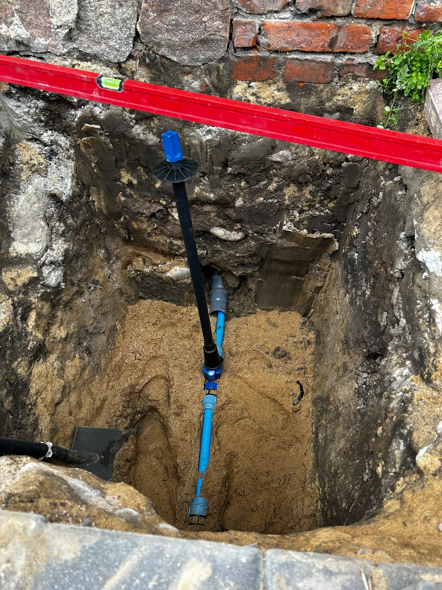 Wykop z piaskiem, w którym widoczne są niebieskie rury instalacji wodnej połączone czarną rurą, obok leży szpadel, a nad wykopem widać poziomicę.
