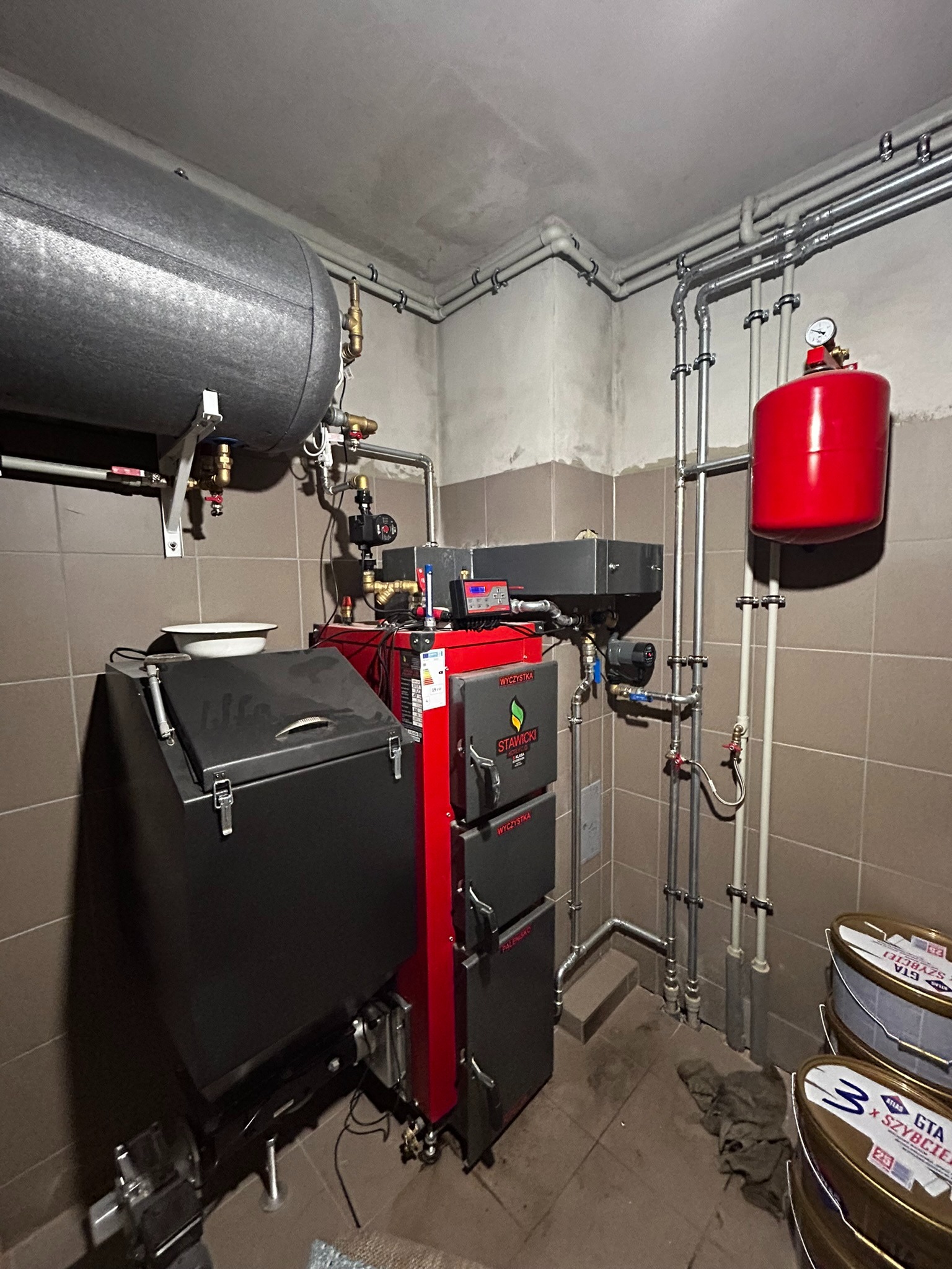 Nowoczesny, czerwony piec na paliwo stałe z podajnikiem, z szarym zasobnikiem na paliwo, zainstalowany w kotłowni z szarymi płytkami, widoczne rury i zbiornik wyrównawczy.