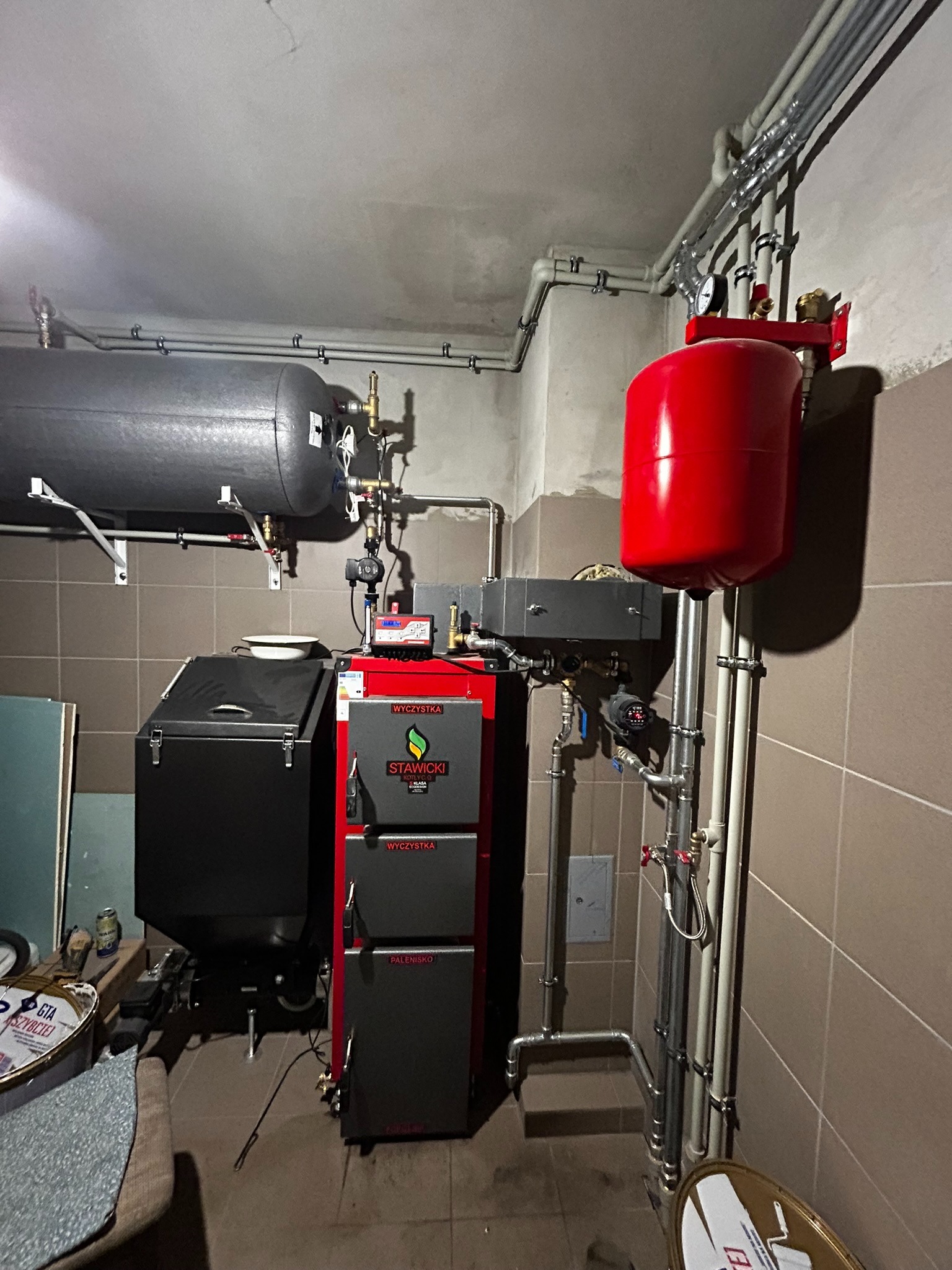 Kotłownia z kotłem na paliwo stałe marki Stawicki, zbiornikiem akumulacyjnym i czerwonym naczyniem wzbiorczym, widoczne rury instalacji grzewczej.