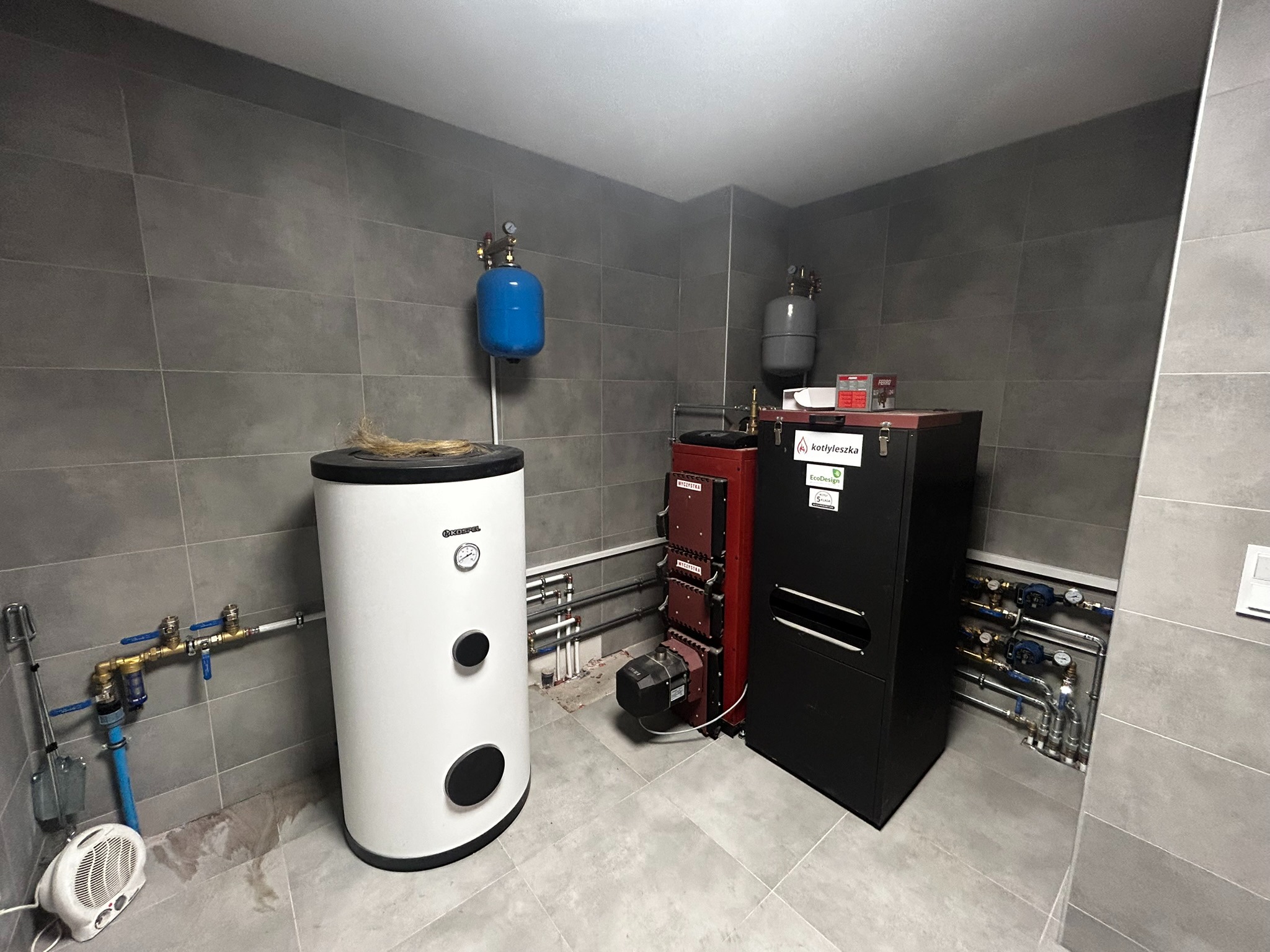 Nowoczesna kotłownia z piecem na paliwo stałe, zasobnikiem ciepłej wody użytkowej i kompletną instalacją hydrauliczną, widoczne rury miedziane i zawory, szare płytki na ścianach i podłodze...