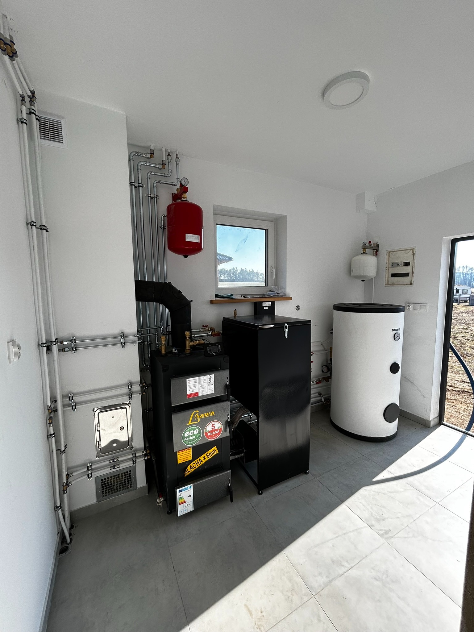 Kotłownia w jasnym pomieszczeniu: czarny piec z etykietami, biały zbiornik akumulacyjny, instalacja rurowa na ścianach i grzejnik wyrównawczy.