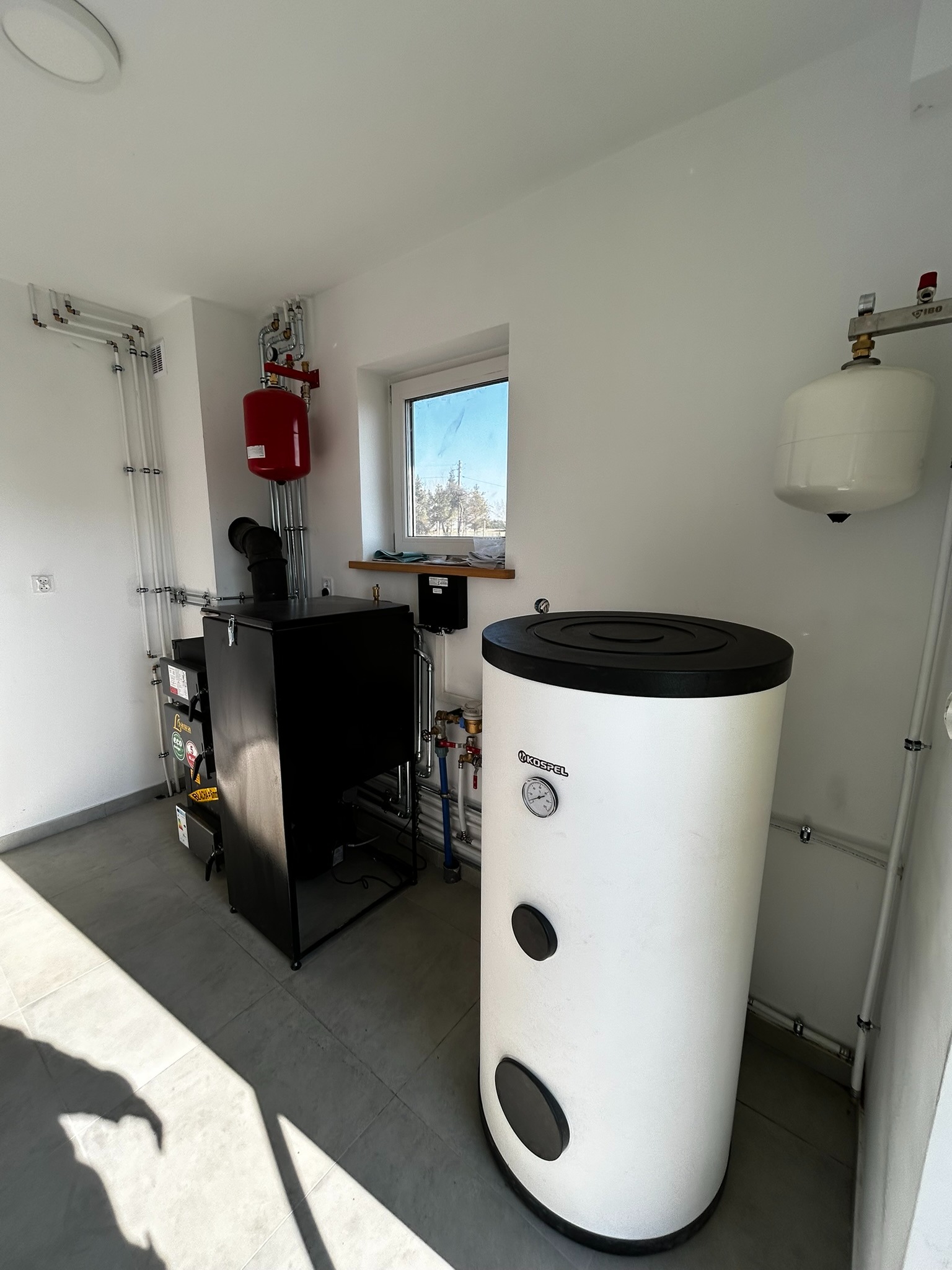 Kotłownia z piecem na paliwo stałe i zasobnikiem ciepłej wody użytkowej marki Kostrzewa, widoczne rury instalacji grzewczej, ekspansomat oraz okno z widokiem na zewnątrz.