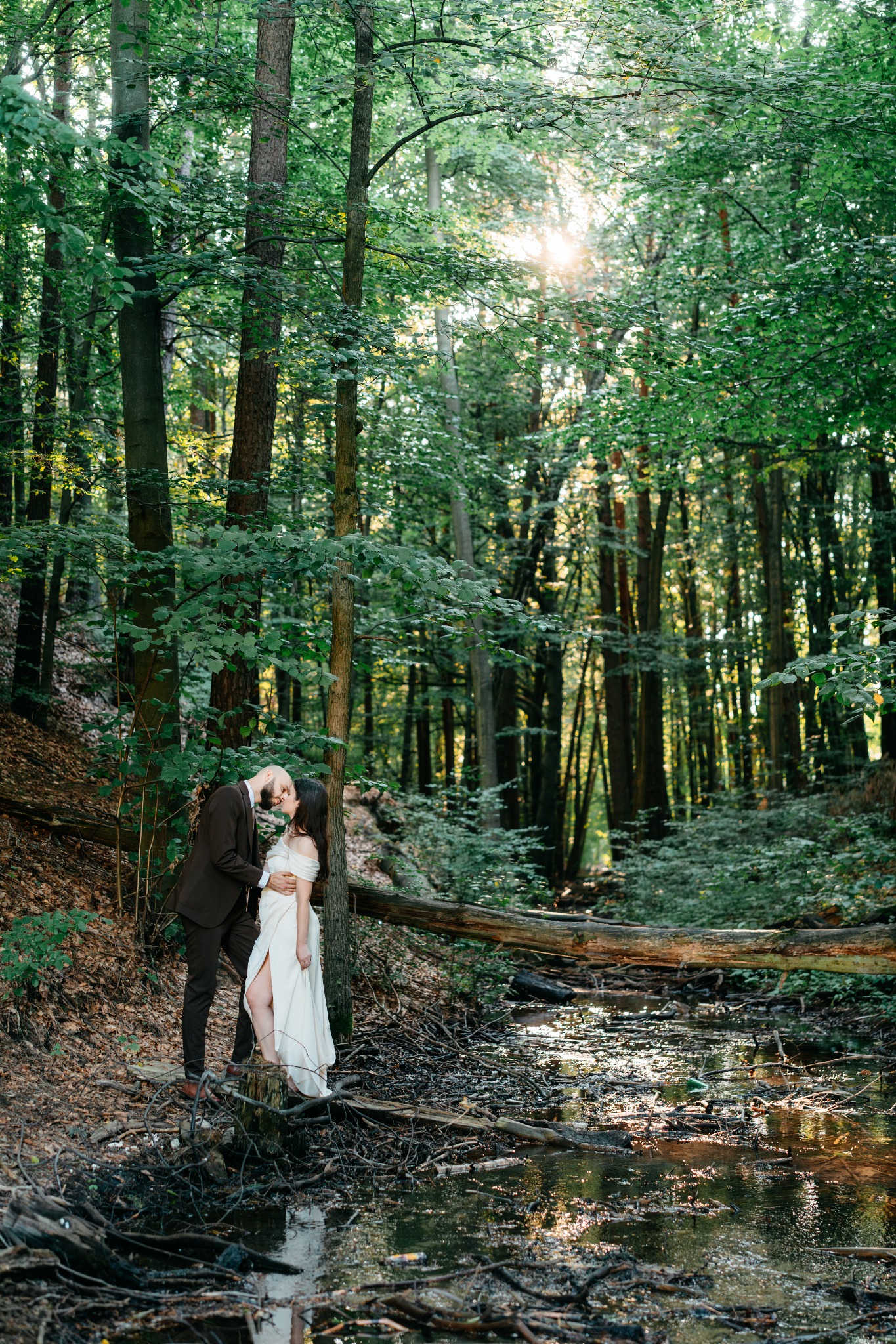 Para młoda całująca się w zalesionym otoczeniu nad małym strumieniem, panna młoda w zwiewnej sukni z rozcięciem, pan młody w ciemnym garniturze.