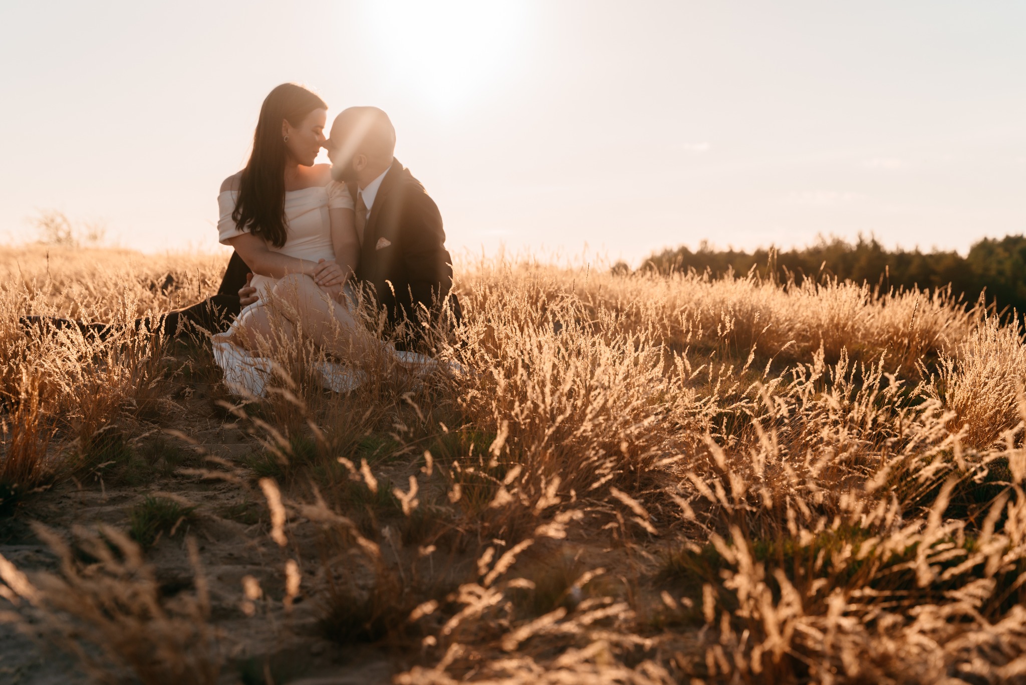Para młoda siedzi w wysokiej, złotej trawie podczas zachodu słońca, delikatnie się dotykając.