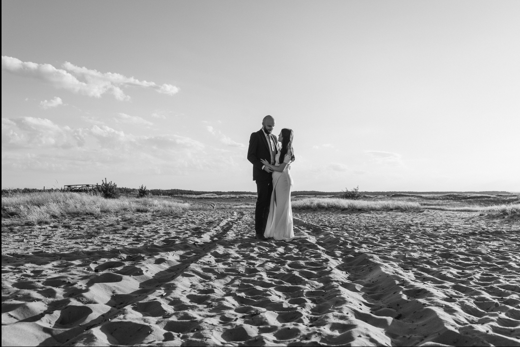 Czarno-białe zdjęcie pary młodej na piaszczystej plaży z wydmami i trawą w tle, silne cienie podkreślają fakturę piasku, para obejmuje się czule, a na horyzoncie widoczne są chmury.
