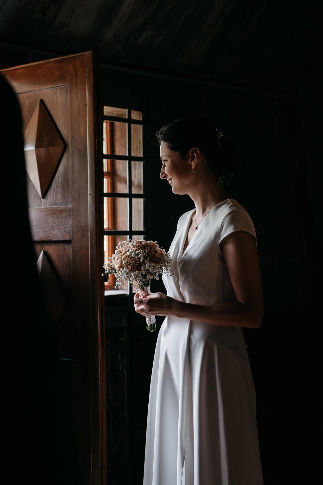 Panna młoda w białej sukni trzyma bukiet, spoglądając przez okno w ciemnym pomieszczeniu z drewnianymi elementami.