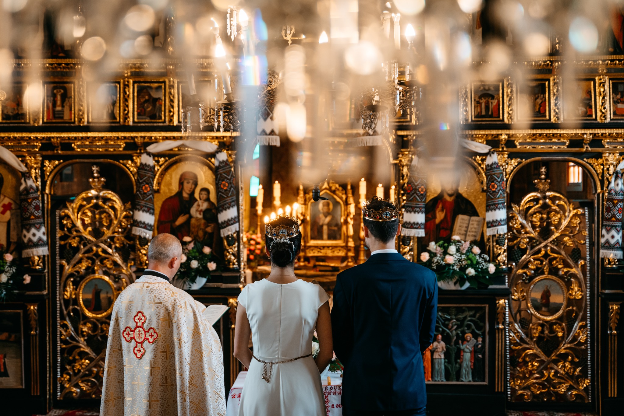 Para młoda w koronach stoi przed ołtarzem podczas ceremonii ślubnej w bogato zdobionym wnętrzu cerkwi, z księdzem w szacie z czerwonym krzyżem po lewej stronie.
