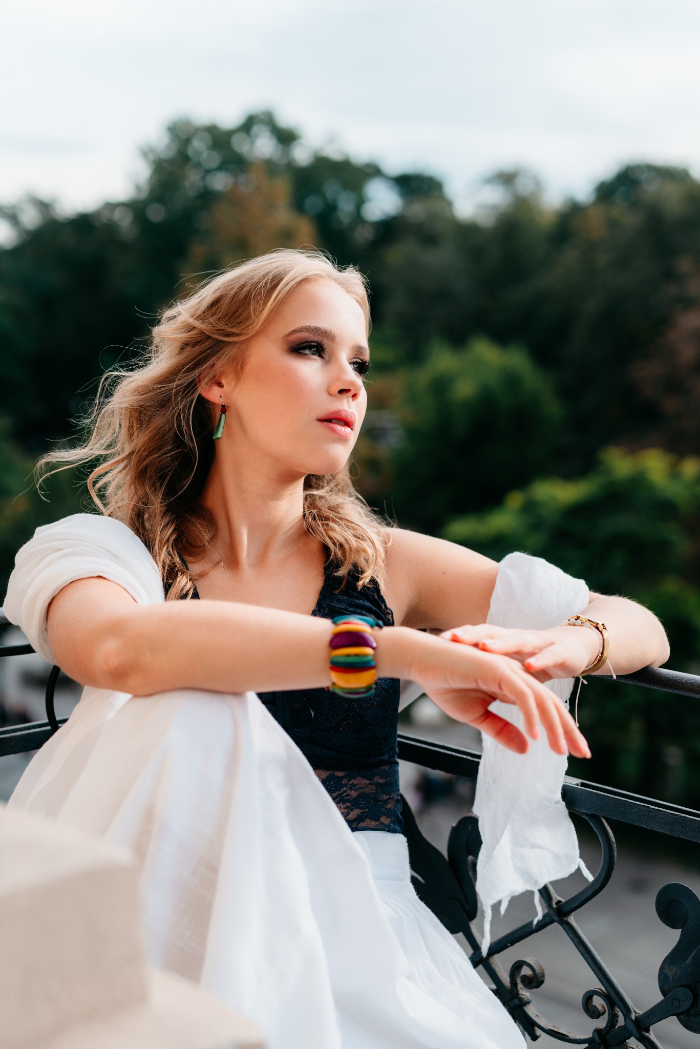 Portret młodej kobiety na balkonie, w czarnej koronkowej bluzce i białej spódnicy, z kolorową bransoletką, zamyślona poza, drzewa w tle.