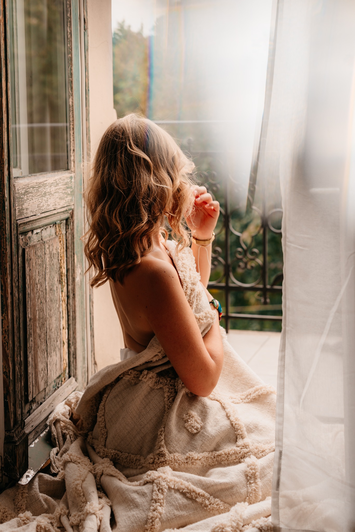 Kobieta z kręconymi włosami, okryta beżowym kocem, siedzi na parapecie obok starego, drewnianego okna z widokiem na balkon i zieleń w tle.