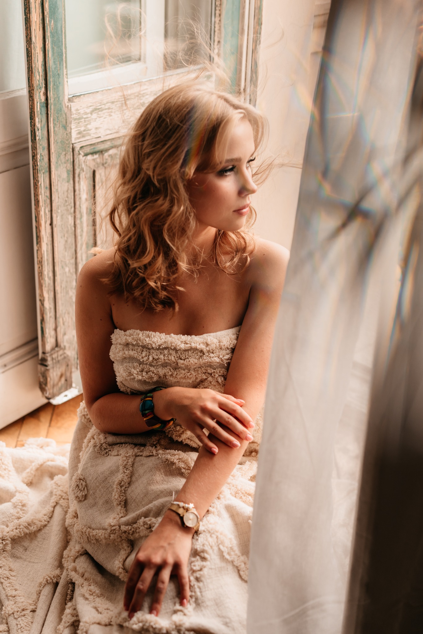 Portret kobiety w zwiewnej sukience, siedzącej przy oknie z efektem tęczy, z widocznymi bransoletkami na nadgarstkach i rozpuszczonymi blond włosami.