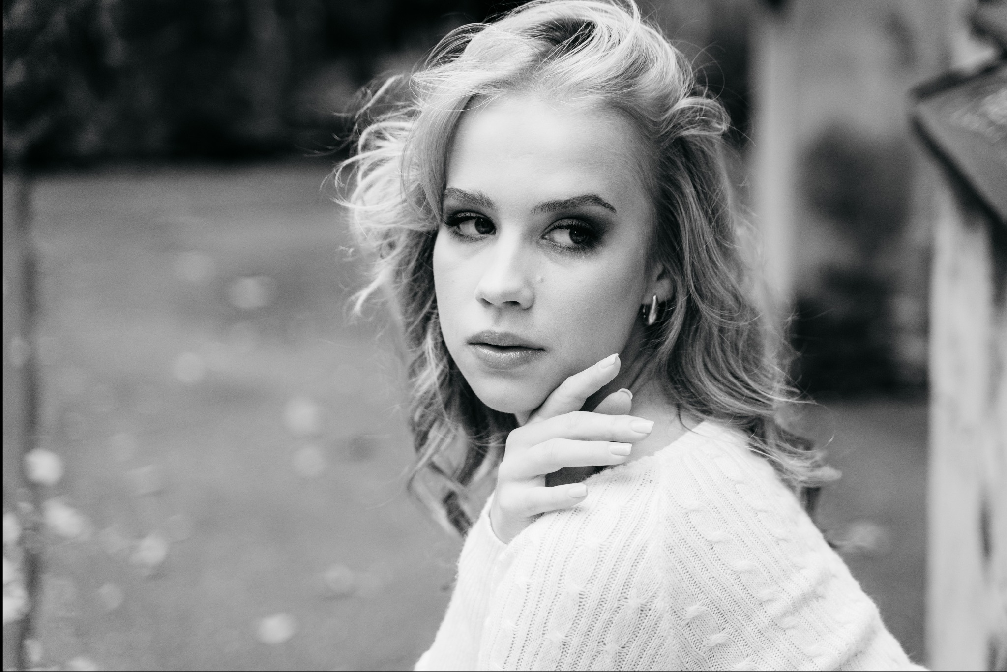 Portret młodej kobiety z blond włosami w swetrze z warkoczami, spoglądającej przez ramię, w czerni i bieli.