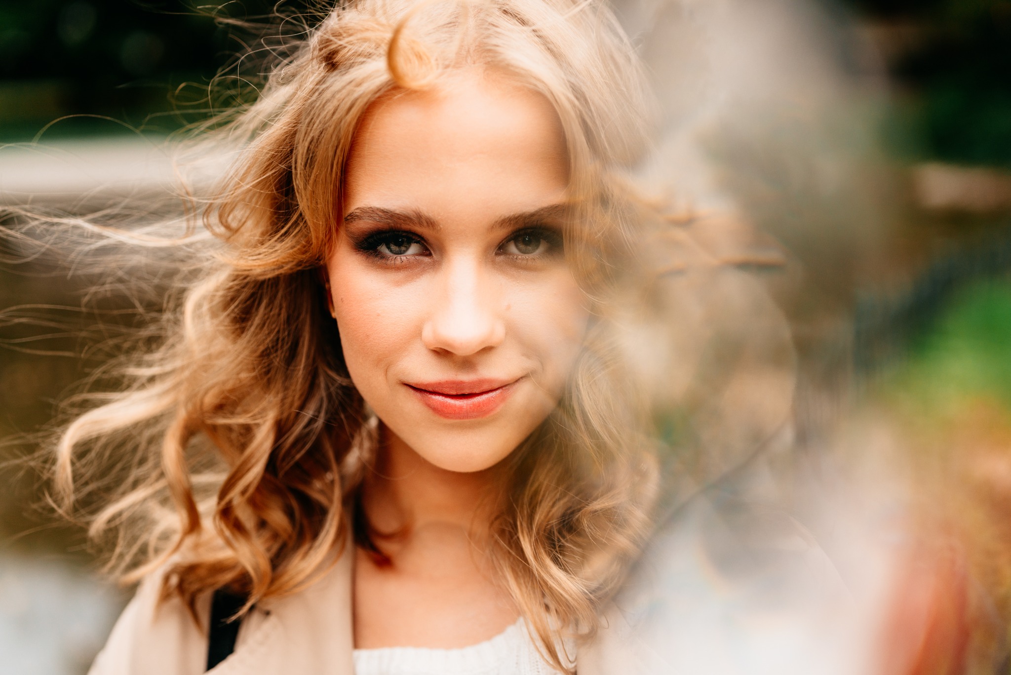 Portret młodej kobiety z blond, kręconymi włosami, lekko uśmiechniętej, z delikatnym makijażem, na rozmytym tle zieleni i brązu.