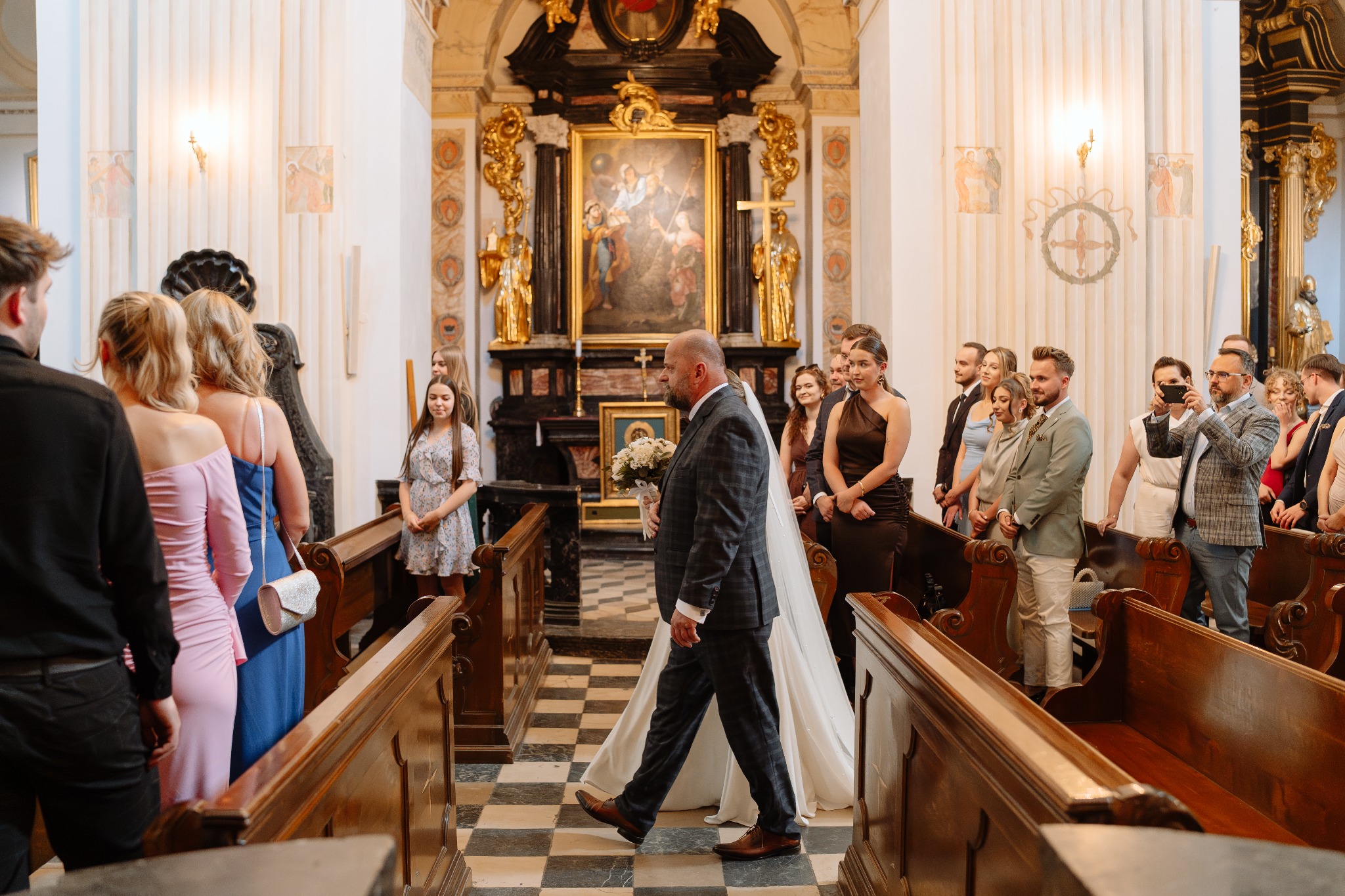 Para młoda idzie w kościele wzdłuż ławek, goście obserwują. Pan młody w garniturze w kratę, panna młoda w białej sukni z welonem. Dekoracje kościelne.