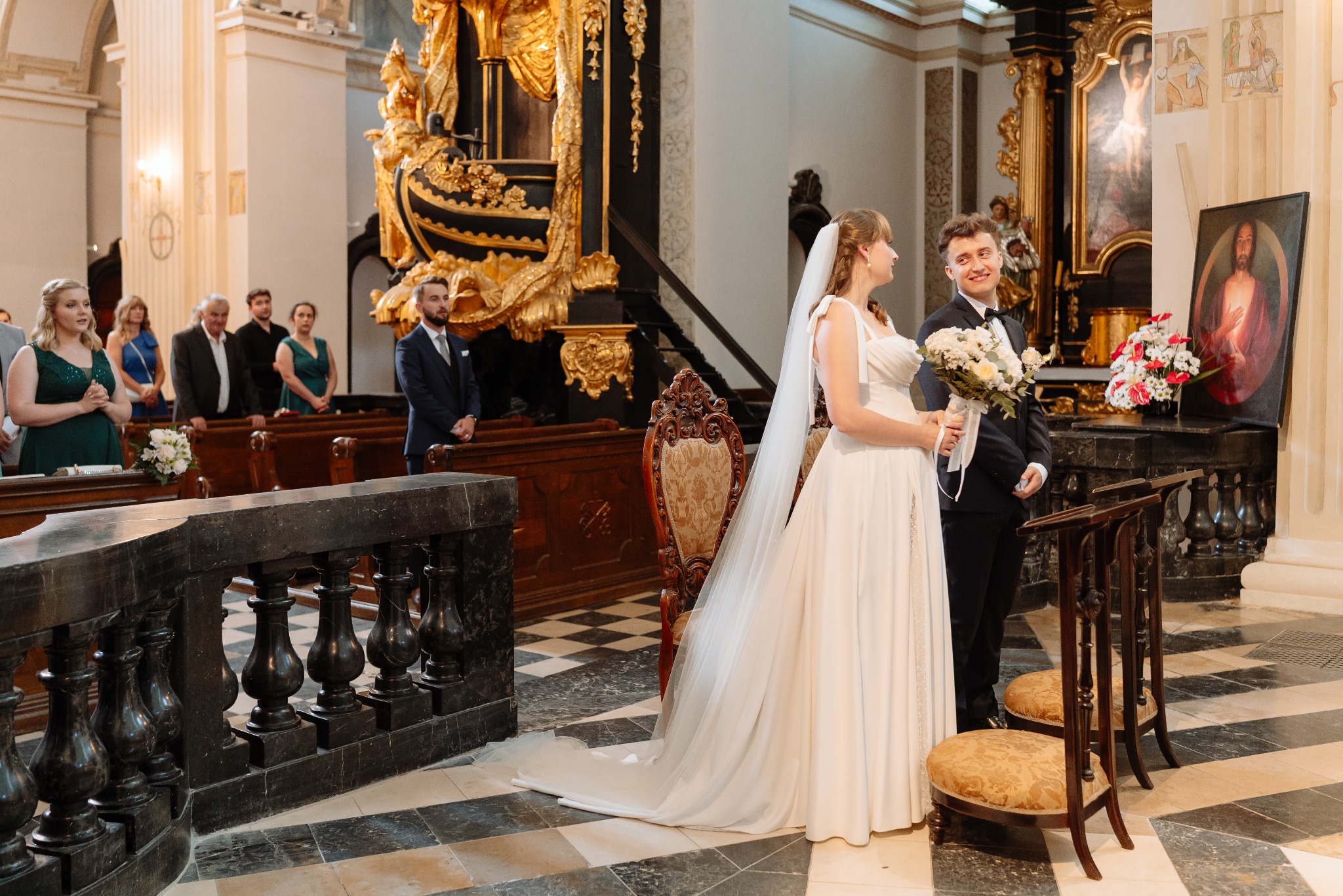 Para młoda w kościele podczas ceremonii ślubnej. Panna młoda w białej sukni, pan młody w garniturze. W tle goście i bogato zdobione wnętrze.