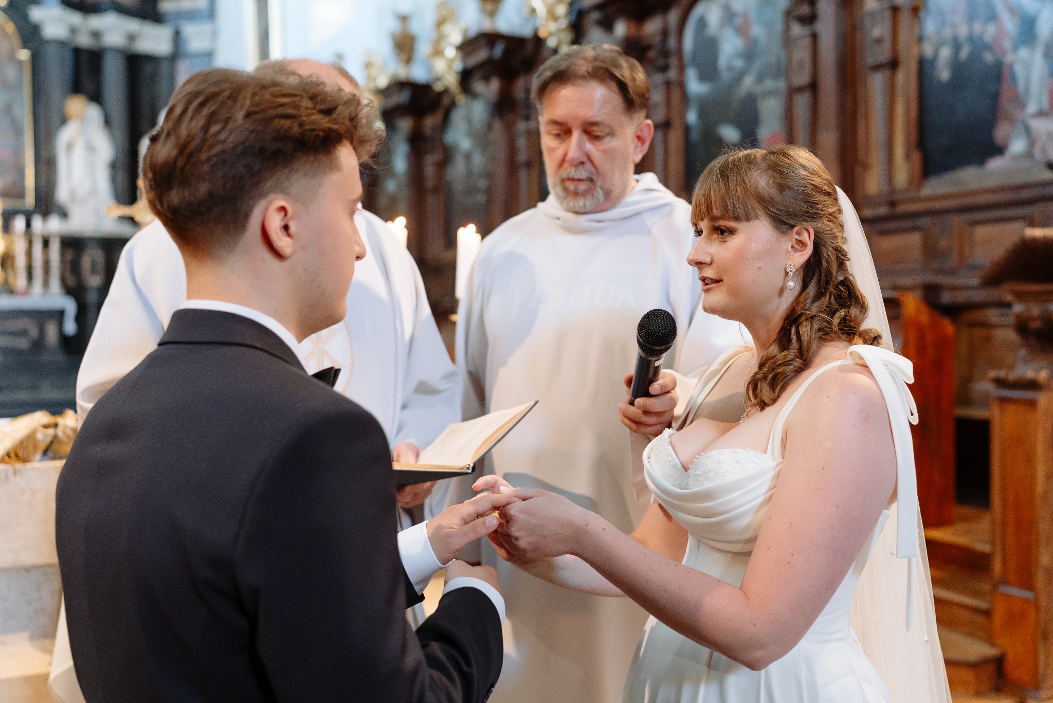 Wymiana obrączek podczas ceremonii ślubnej w kościele. Para młoda stoi przed księdzem, który trzyma mikrofon i księgę. W tle widać ołtarz i świece.