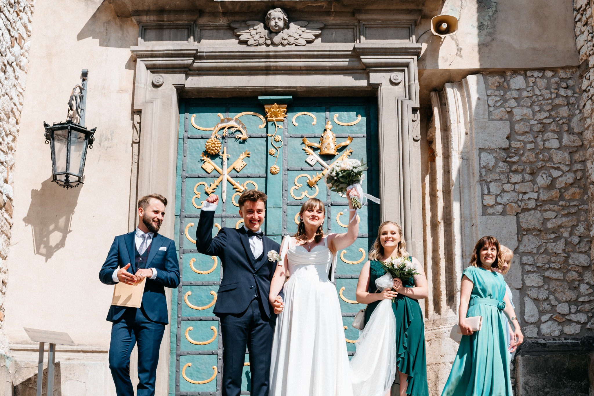 Uśmiechnięta para młoda z gośćmi weselnymi świętuje przed zdobionymi, turkusowymi drzwiami z herbami. W tle kamienna ściana i latarnia.