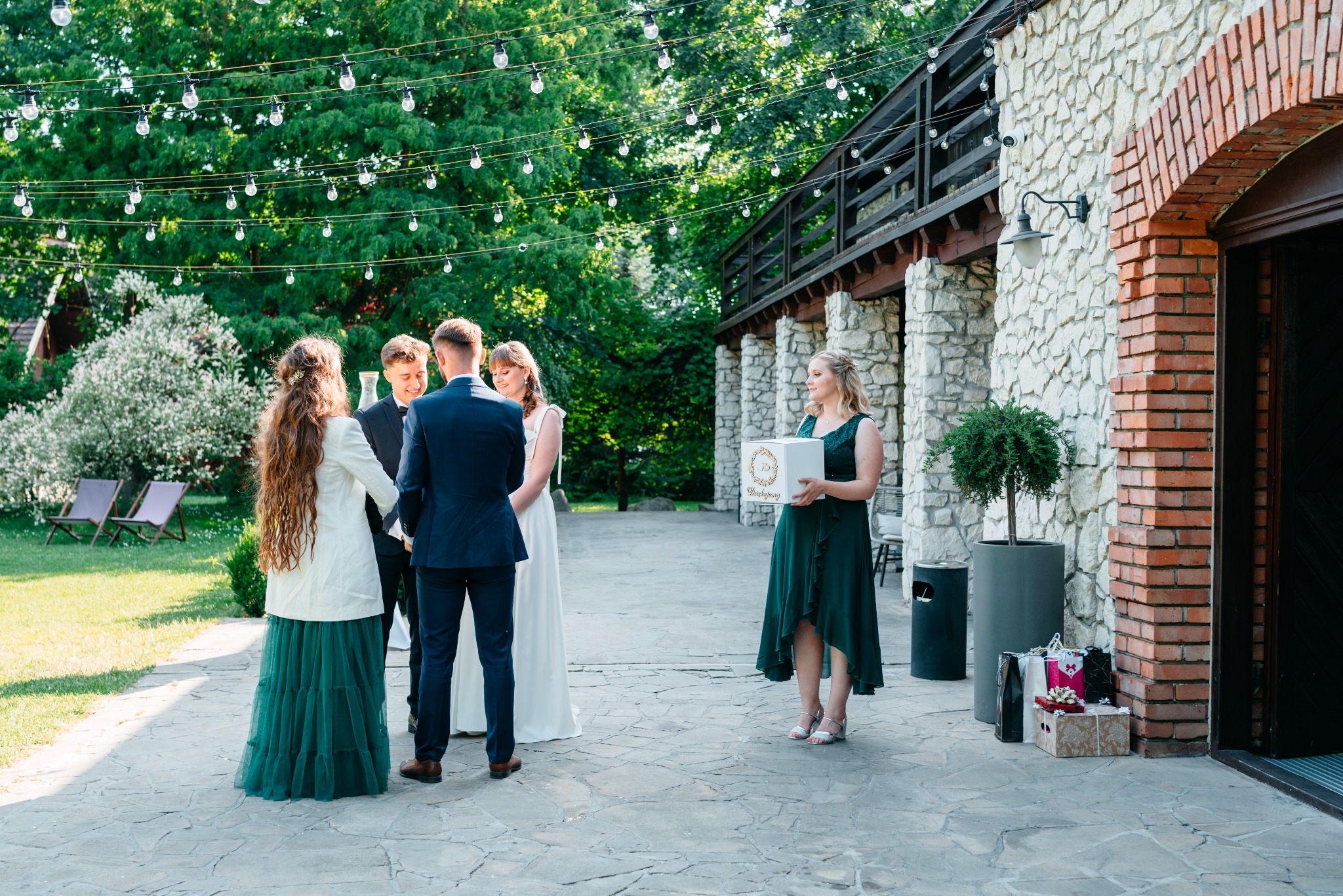 Uroczystość ślubna w plenerze: Panna młoda w białej sukni, goście w eleganckich strojach, dekoracje świetlne, kamienna architektura w tle.