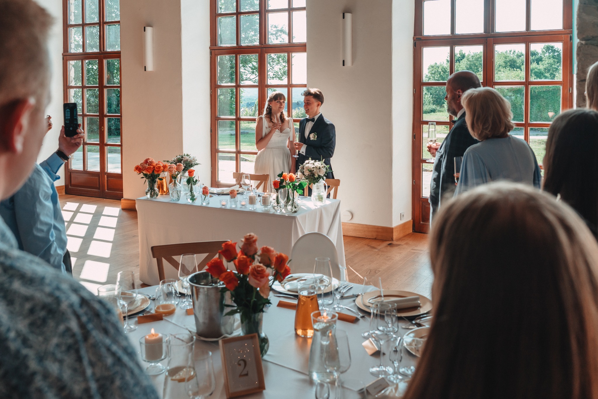 Para młoda wznosi toast podczas przyjęcia weselnego w przestronnej sali z dużymi oknami. Dekoracje kwiatowe, eleganckie nakrycia stołów, goście w tle.