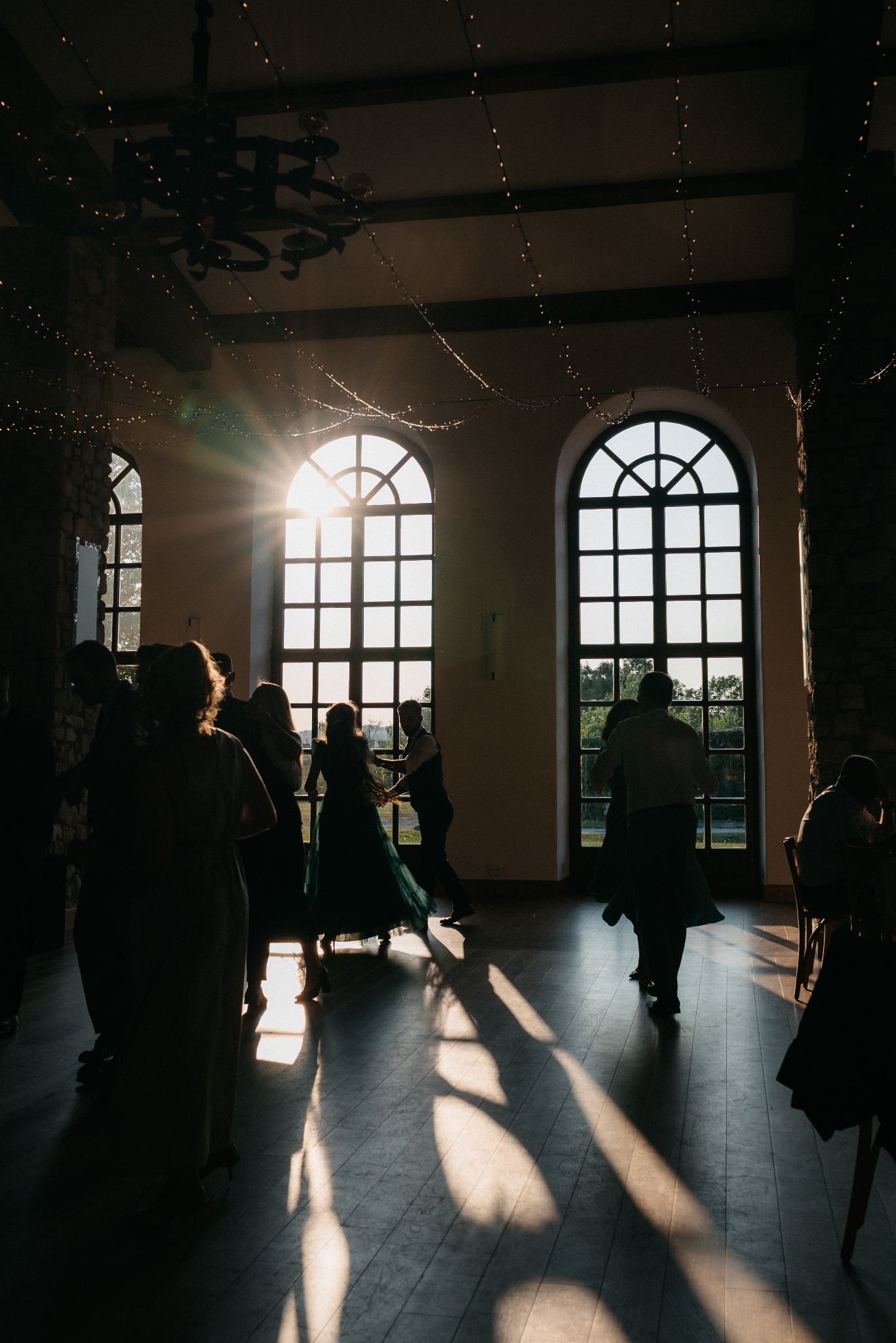 Sylwetki tańczących gości weselnych w sali z dużymi oknami i dekoracyjnym oświetleniem. Słońce wpada przez okna, tworząc długie cienie na parkiecie.