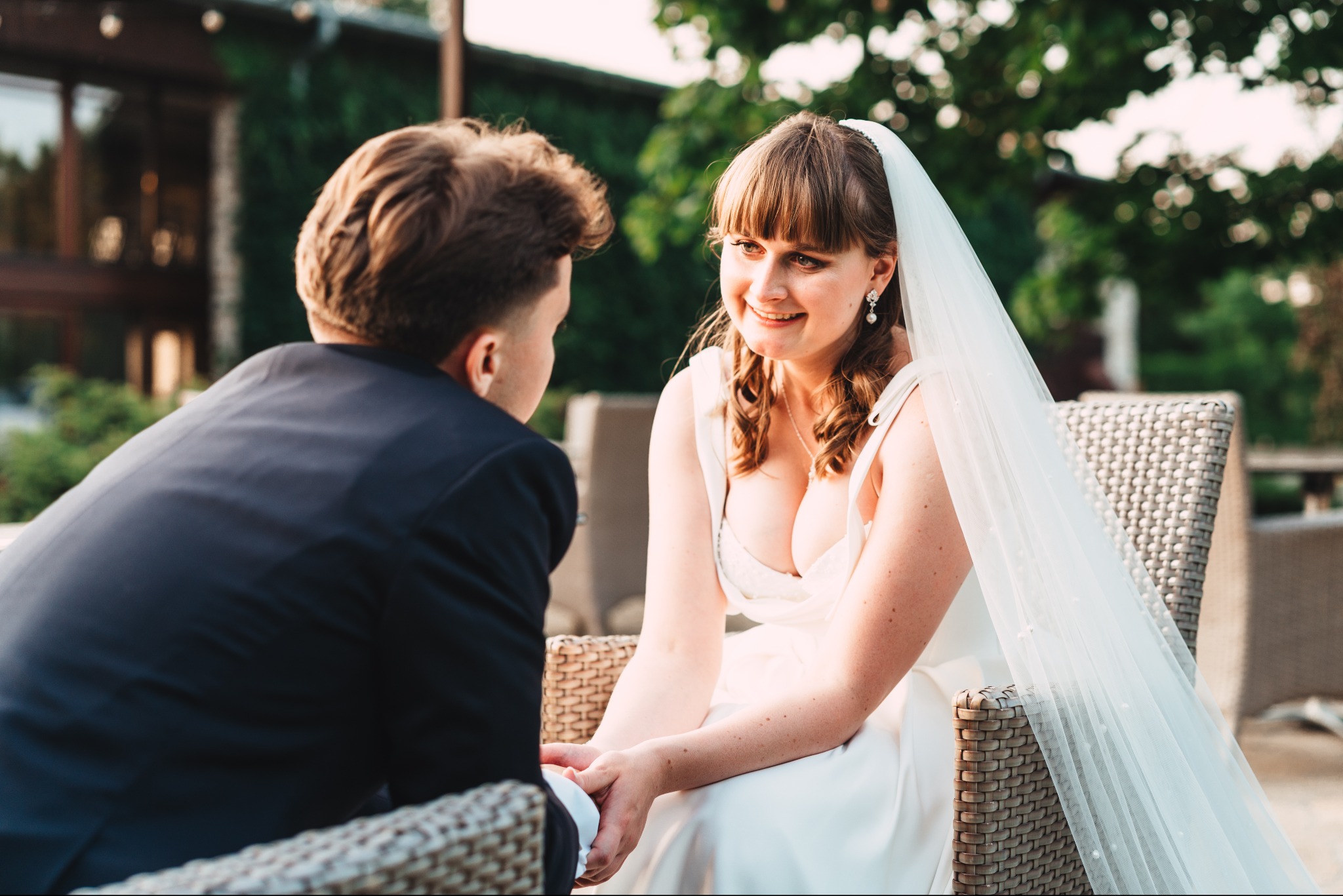 Uśmiechnięta panna młoda w białej sukni i welonie, siedzi naprzeciwko pana młodego trzymając go za rękę, na tle zieleni i zabudowań.
