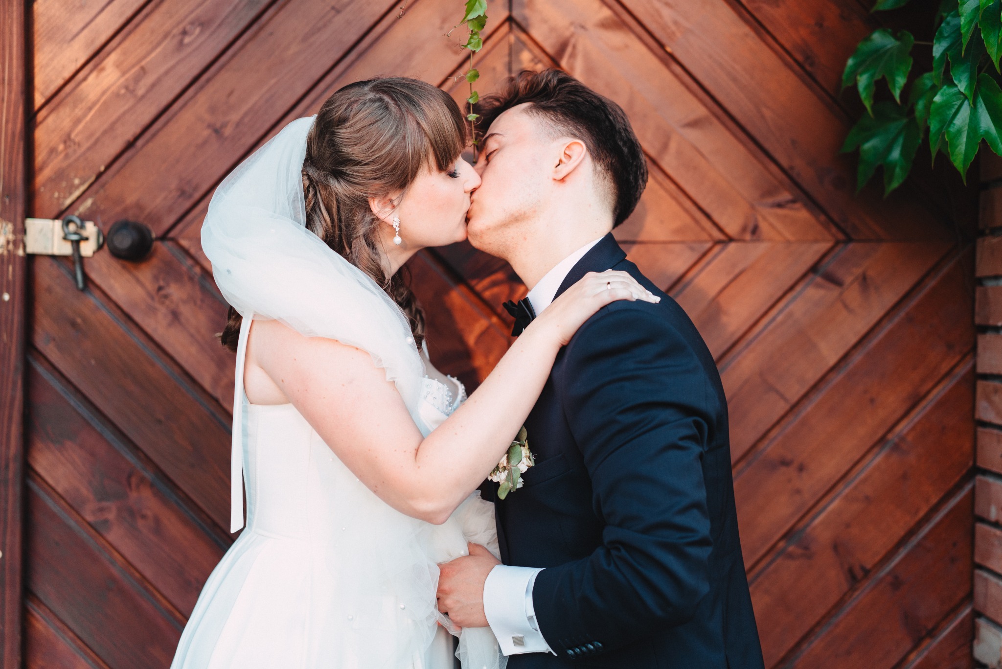 Pocałunek pary młodej na tle drewnianych drzwi z roślinnym akcentem. Panna młoda w białej sukni i welonie, pan młody w granatowym garniturze z muchą. Romantyczna fotografia ślubna.