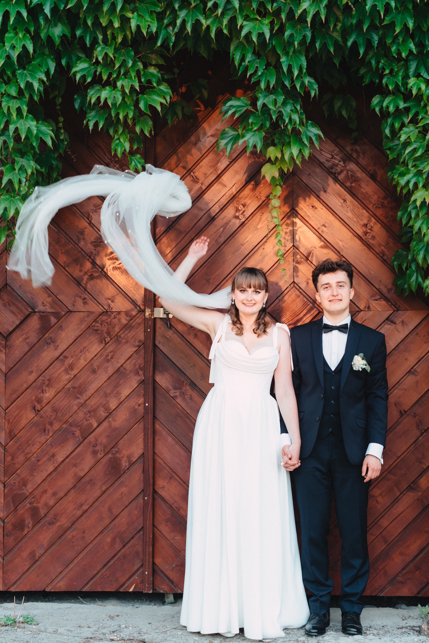 Para młoda trzymająca się za ręce na tle drewnianych wrót porośniętych bluszczem, panna młoda unosi welon. Ujęcie z sesji ślubnej.