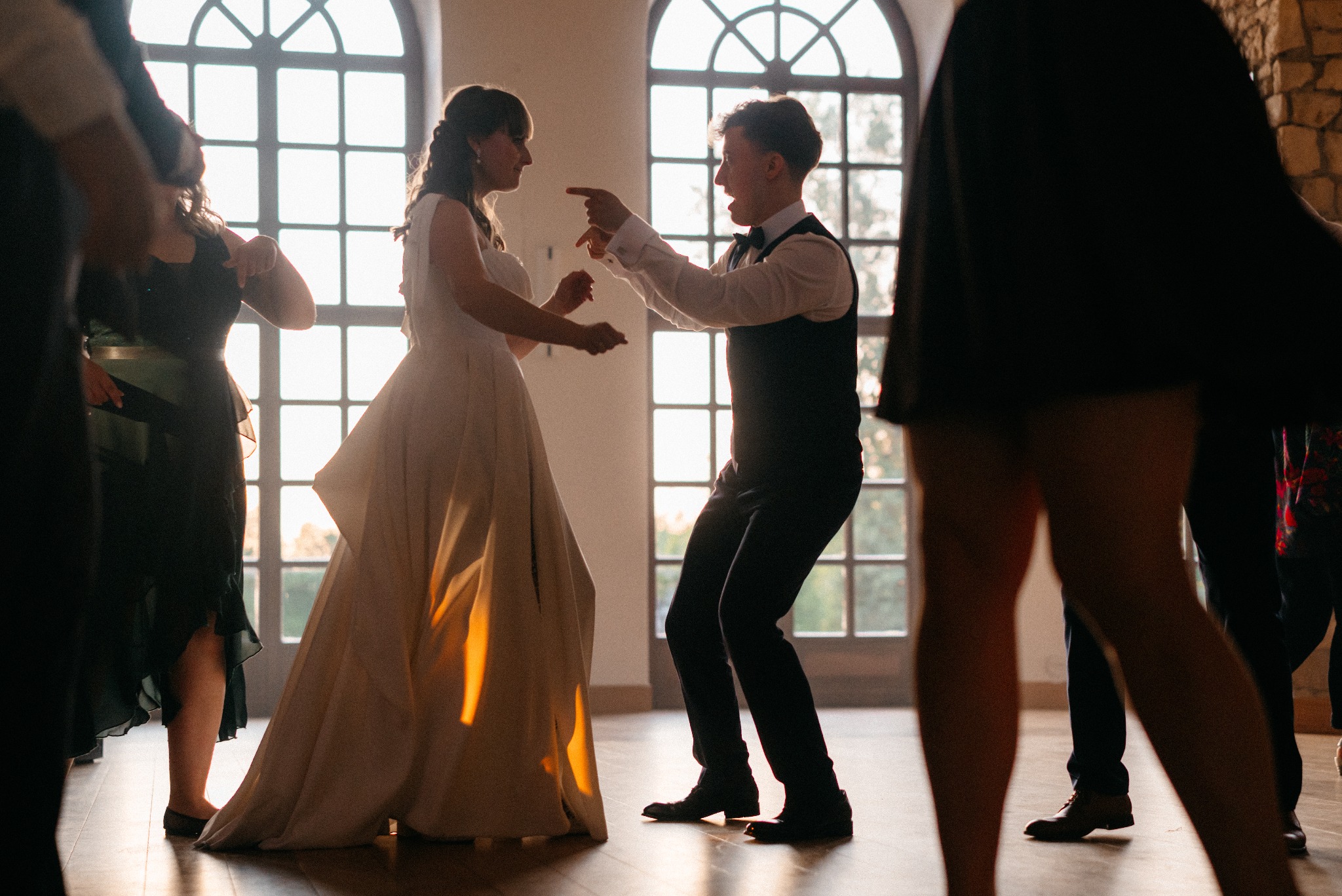 Para młoda podczas żywiołowego tańca na parkiecie, otoczona gośćmi weselnymi. Duże okno w tle, ciepłe światło zachodzącego słońca.