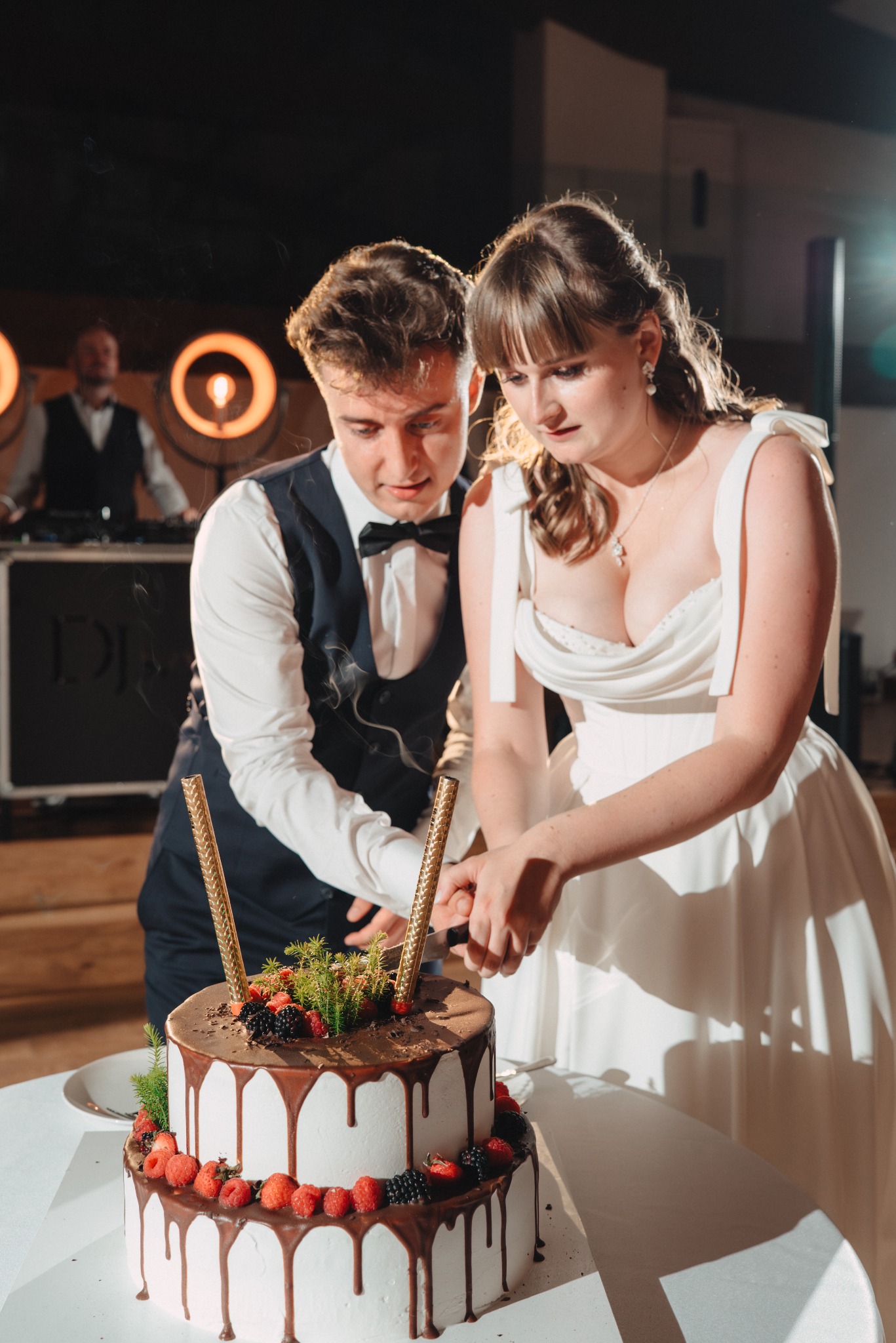Uśmiechnięta para młoda kroi tort weselny, w tle DJ na tle świateł. Dekoracja tortu z owocami i świecami. Ujęcie z bliska na uroczystości weselnej.