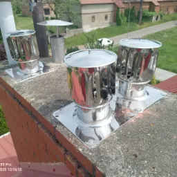 Błyszczące, metalowe nasady kominowe, typu turbowent, zamontowane na ceglanym kominie z szarą, betonową czapą, na tle zieleni i budynków. Widok z góry.