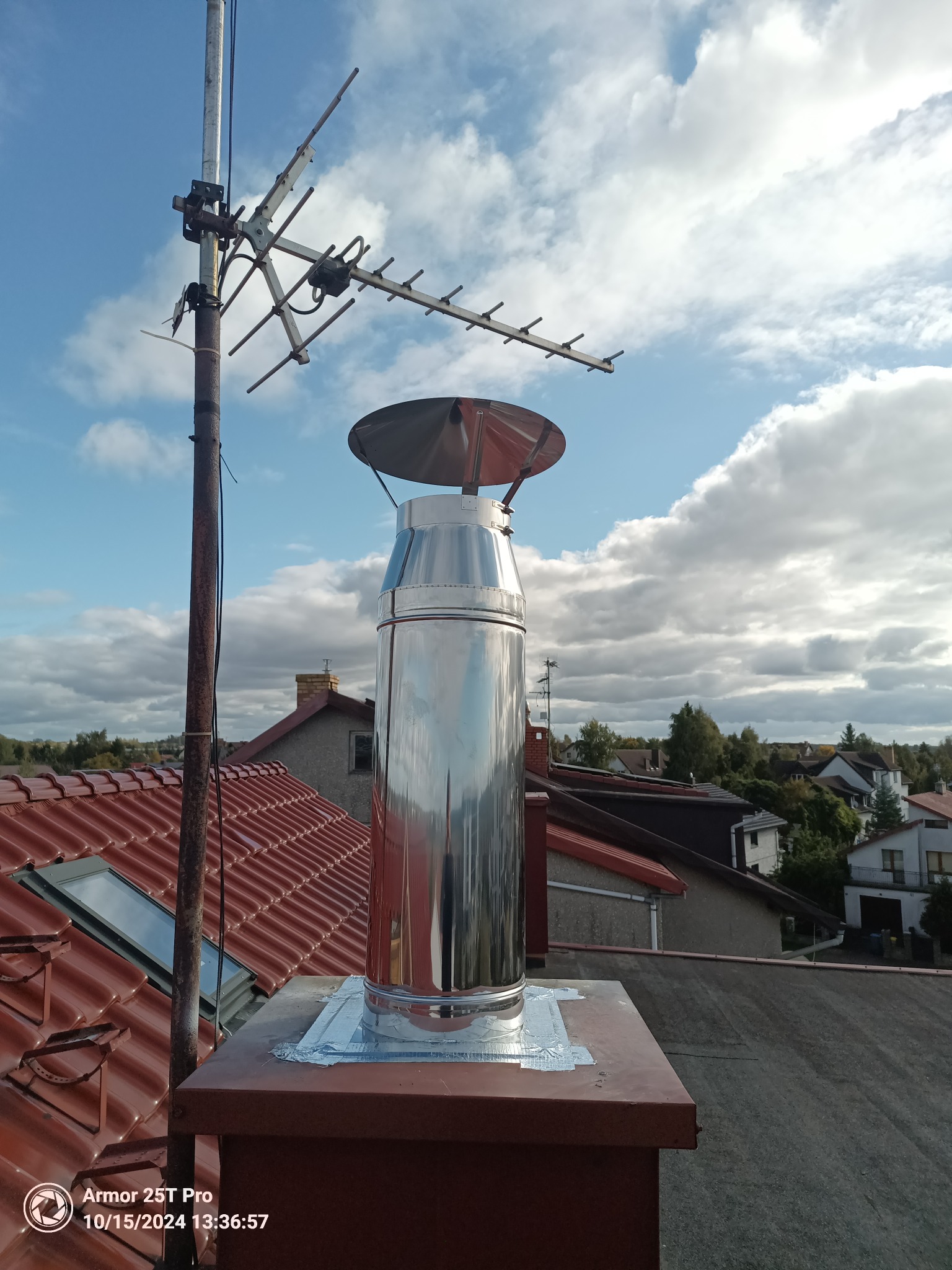 Błyszczący, nowy wkład kominowy ze stali nierdzewnej, zamontowany na kominie z brązowej cegły, zabezpieczony taśmą aluminiową, na tle czerwonego dachu i nieba z chmurami, obok maszt antenowy.